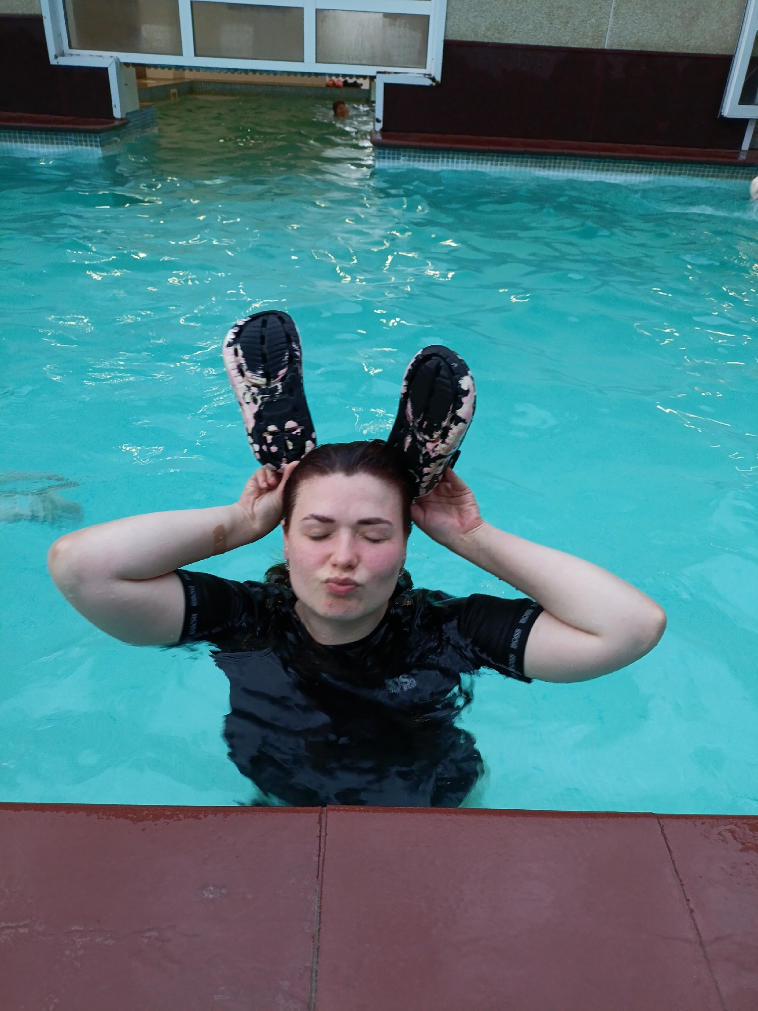 The image depicts an adult female engaged in a playful activity within an indoor swimming pool, specified as being in the city of Chaikovsky, Russia. The woman, who appears to be in her 20s to 30s with dark hair, is submerged approximately chest-deep in clear, turquoise water. She is wearing a black short-sleeved swim top, possibly with a "BOSS" logo visible on the left sleeve. Her eyes are closed, and her lips are pursed in a pronounced 'kissing' or 'fish' pose, indicating a deliberate humorous expression. With both hands, she holds a black and pink/white patterned sandal above each side of her head, positioning them like animal ears with the soles facing outwards. A small, light-colored bandage or dressing is visible on her right forearm.
The pool area features a reddish-brown tiled or composite material forming the immediate surround of the pool. In the background, another individual's head is partially visible above the water in what appears to be a separate or deeper section of the pool. The background architecture includes light-colored tiled walls topped by white-framed windows, with a darker reddish-brown strip beneath the windows, suggesting an indoor facility such as a public swimming complex or a hotel pool. The water exhibits minor surface ripples.