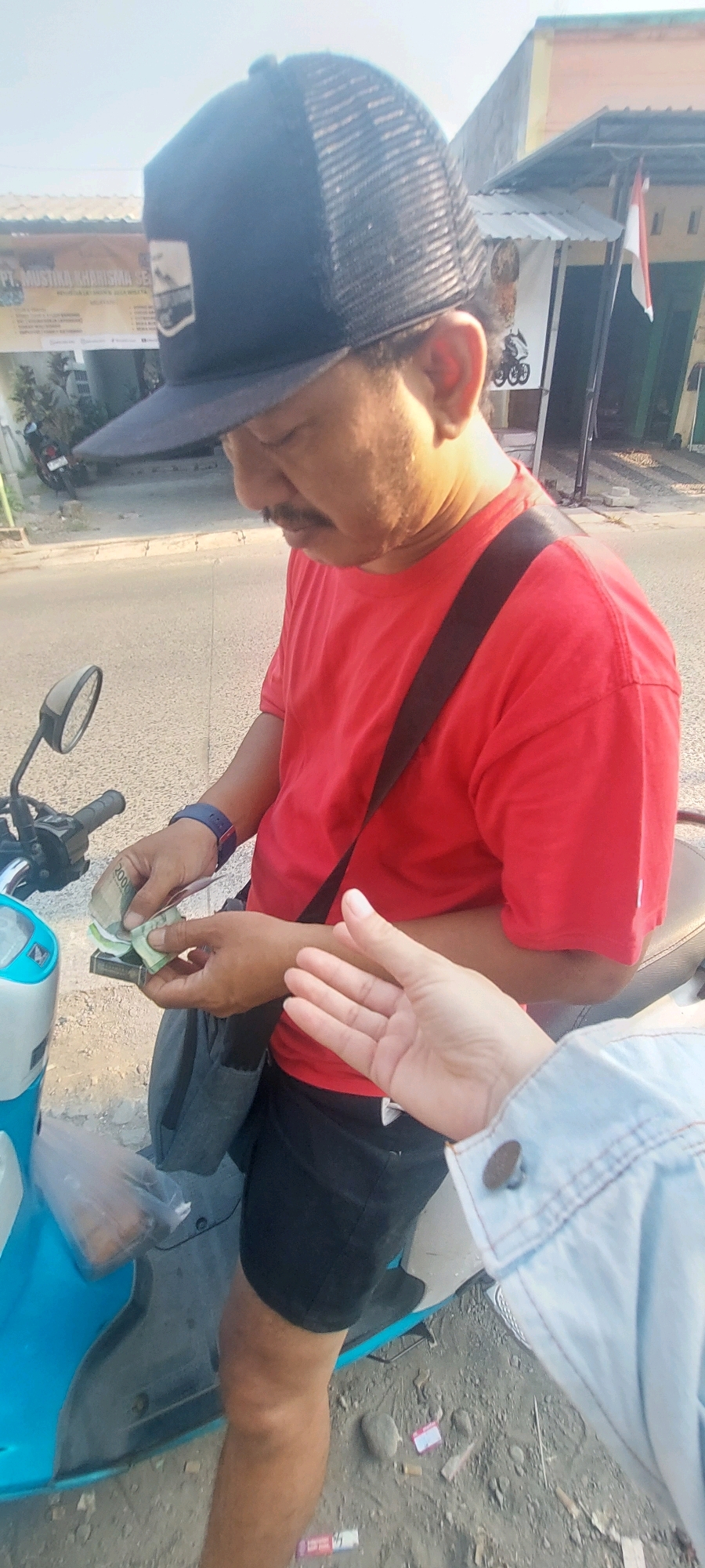 A man wearing a red t-shirt, black shorts, and a black mesh-backed baseball cap stands next to a light blue and white motorcycle. He is holding a wad of Indonesian Rupiah banknotes in his left hand and appears to be counting them. A dark grey bag is slung across his chest, and a blue watch is visible on his left wrist. A second person's hand, wearing a light blue denim jacket, is visible in the lower right of the frame, reaching towards the man. The scene is outdoors, with a paved road in the background and a building with an Indonesian flag visible. The ground is composed of dirt and gravel, with some discarded cigarette butts scattered about. The lighting suggests it is daytime. This appears to be a street scene in Semarang, Indonesia, possibly a transaction taking place on the roadside.
