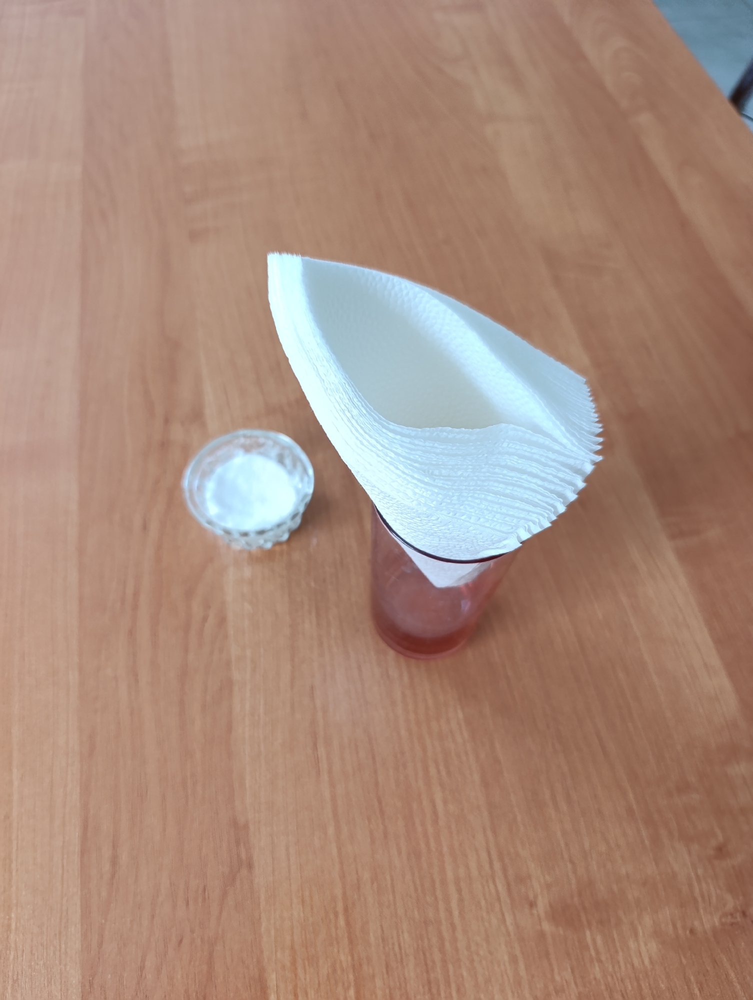 An overhead shot on a wooden table shows a clear glass with a red liquid, a stack of white paper filters inside the glass, and a small glass bowl filled with white powder to the left of the glass. The table has a light brown wood grain pattern. The Dubăsari town council, Moldova is not depicted in this image.