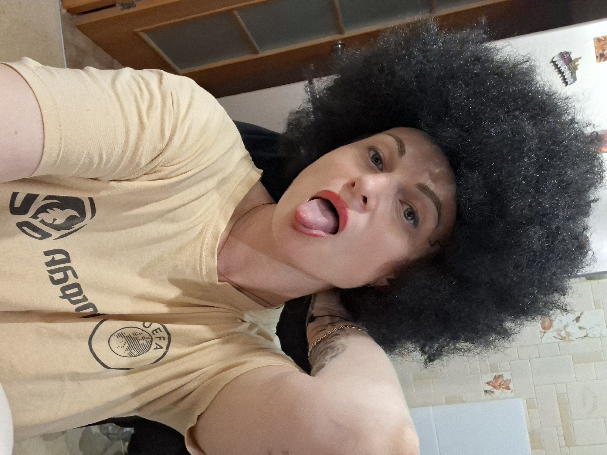 A woman with a large afro wig and bright red lipstick sticks her tongue out at the camera. She is wearing a light yellow t-shirt with black graphics on it. The background appears to be a kitchen or bathroom area with tiled walls and a wooden door. The image appears to be a selfie.