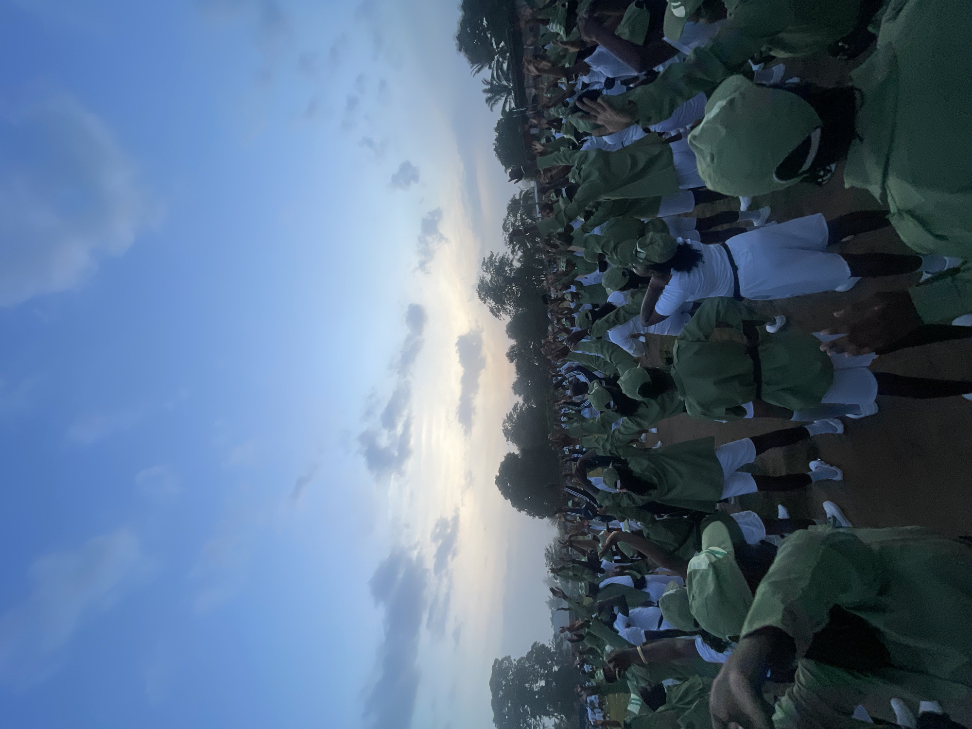 An outdoor, ground-level, full shot captures a large group of young adults, presumably participants in a National Youth Service Corps (NYSC) program, engaged in physical activity under a pale blue, cloudy sky at dawn. The individuals, dressed in matching green shirts, white shorts, and green caps, are spread across a dirt field bordered by trees and a distant structure with a fence. Their arms are raised in unison, suggesting a coordinated exercise or dance. The scene is one of communal activity and discipline, characteristic of a structured training environment. The low light suggests early morning.