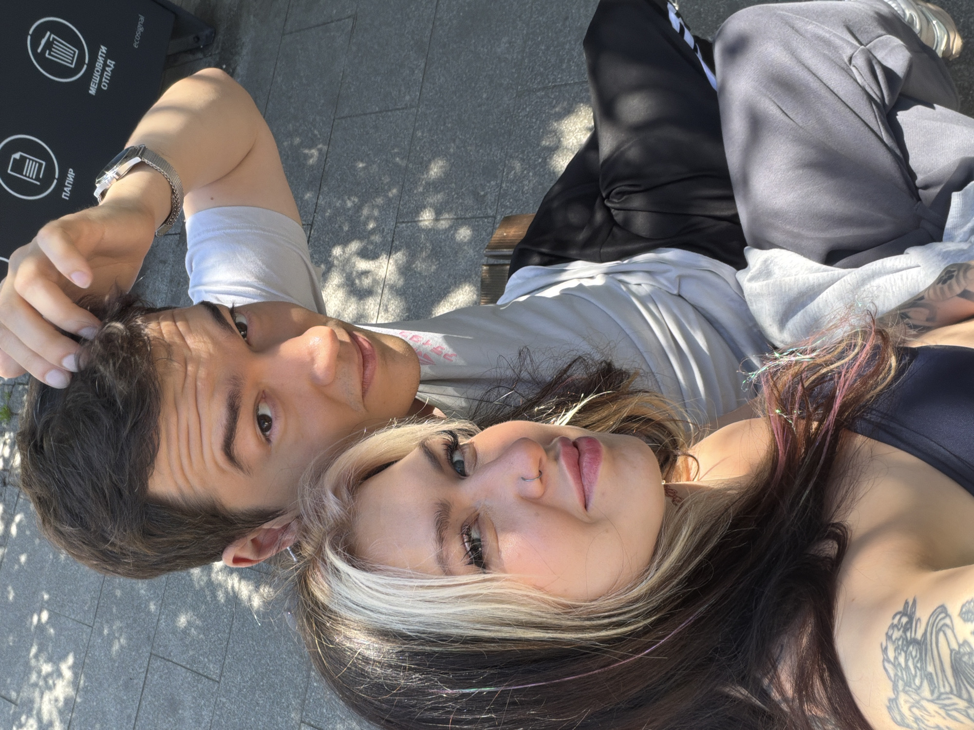 Two people, a man and a woman, are lying down together in an outdoor setting. The man is wearing a light-colored t-shirt and has a watch on his left wrist. The woman has dark hair with blonde streaks and a tattoo on her chest/shoulder area. They appear to be on a bench or flat surface with a gray tiled floor. A recycling bin is visible in the background to the left. The location is Palilula Urban Municipality, Serbia.
