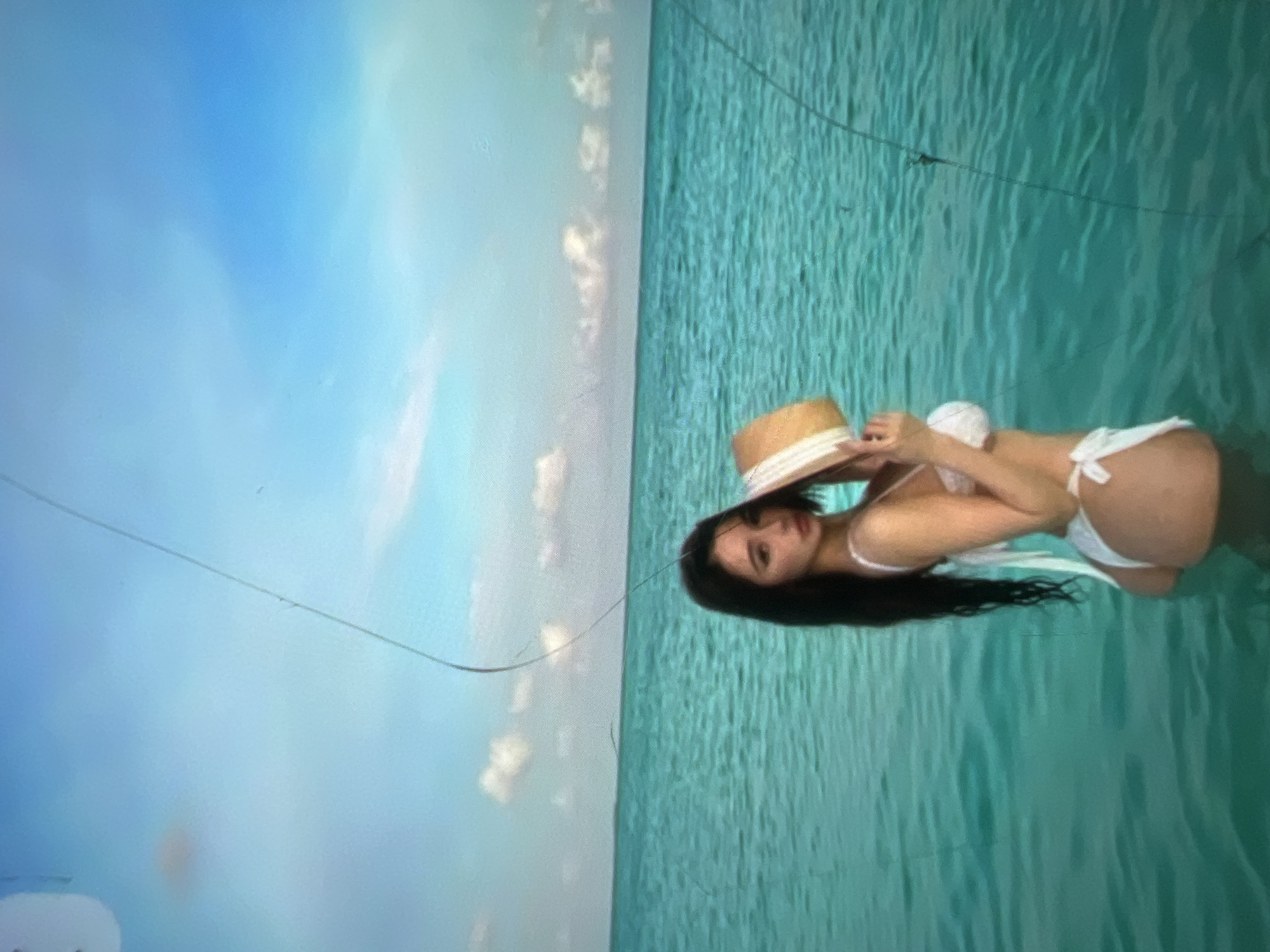 A woman wearing a white bikini and holding a straw hat stands in clear turquoise water. She has long dark hair and is looking over her shoulder at the camera. The background consists of the ocean and a bright blue sky with scattered white clouds. There are faint scratches or marks on the image itself. The location is described as Kara-Dıykan, Kyrgyzstan, though the visual elements do not specifically suggest this location.