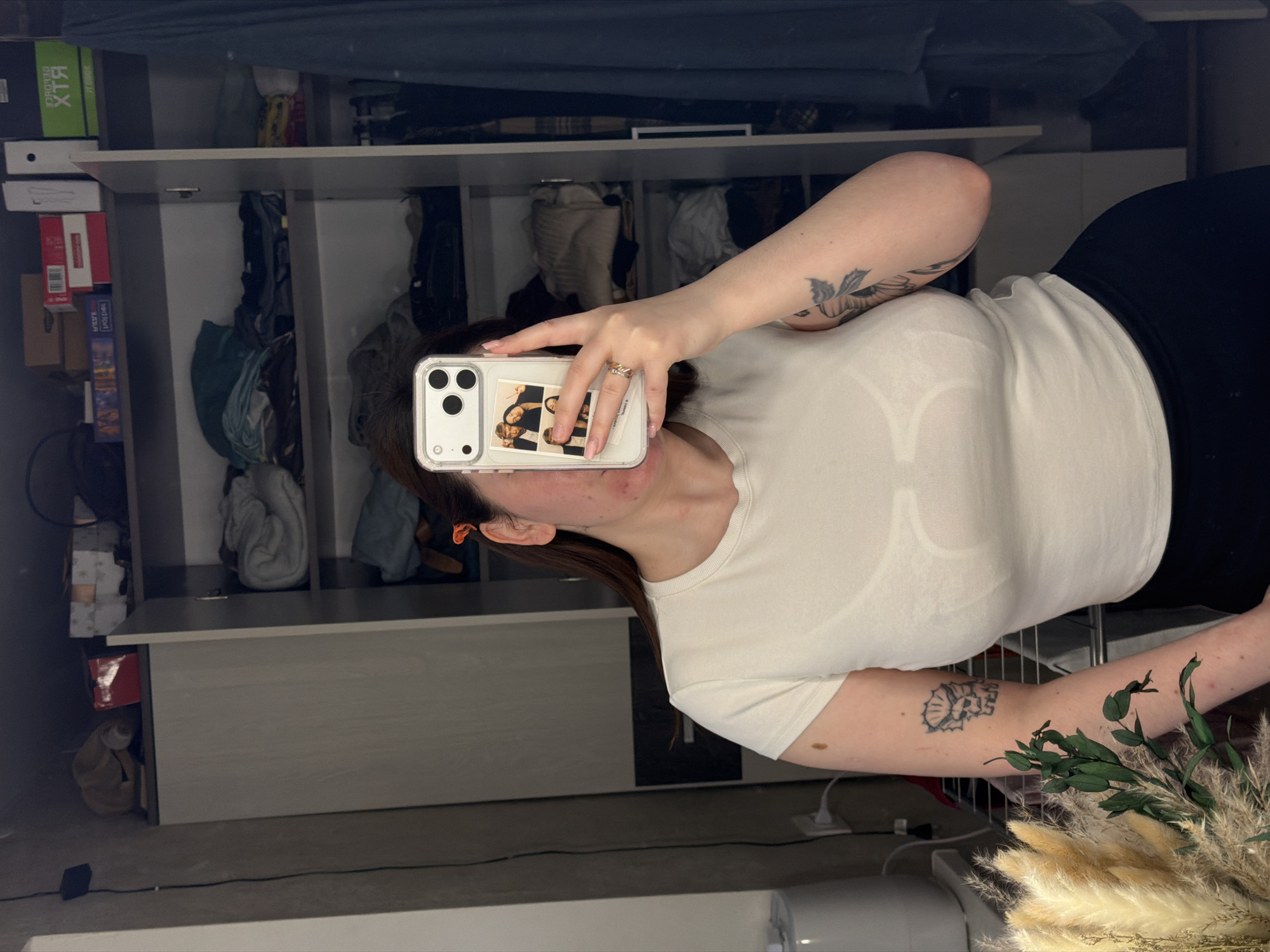 A person is holding a white smartphone up to take a selfie in front of a mirrored closet or wardrobe, seemingly indoors. The person wears a white t-shirt and black leggings. The phone case has several small photos tucked into it. There is a ring on the person's finger on the hand holding the phone. The person has tattoos on their arms. A bouquet of pampas grass is visible in the lower right corner of the image.
The closet or wardrobe behind the person is light gray with various shelves and compartments. Items, likely clothes and boxes, are visible within the closet. A box of RTX computer hardware is seen on one shelf. The environment appears to be a room in a home. There is no evidence of a location of Chaikovsky, Russia.