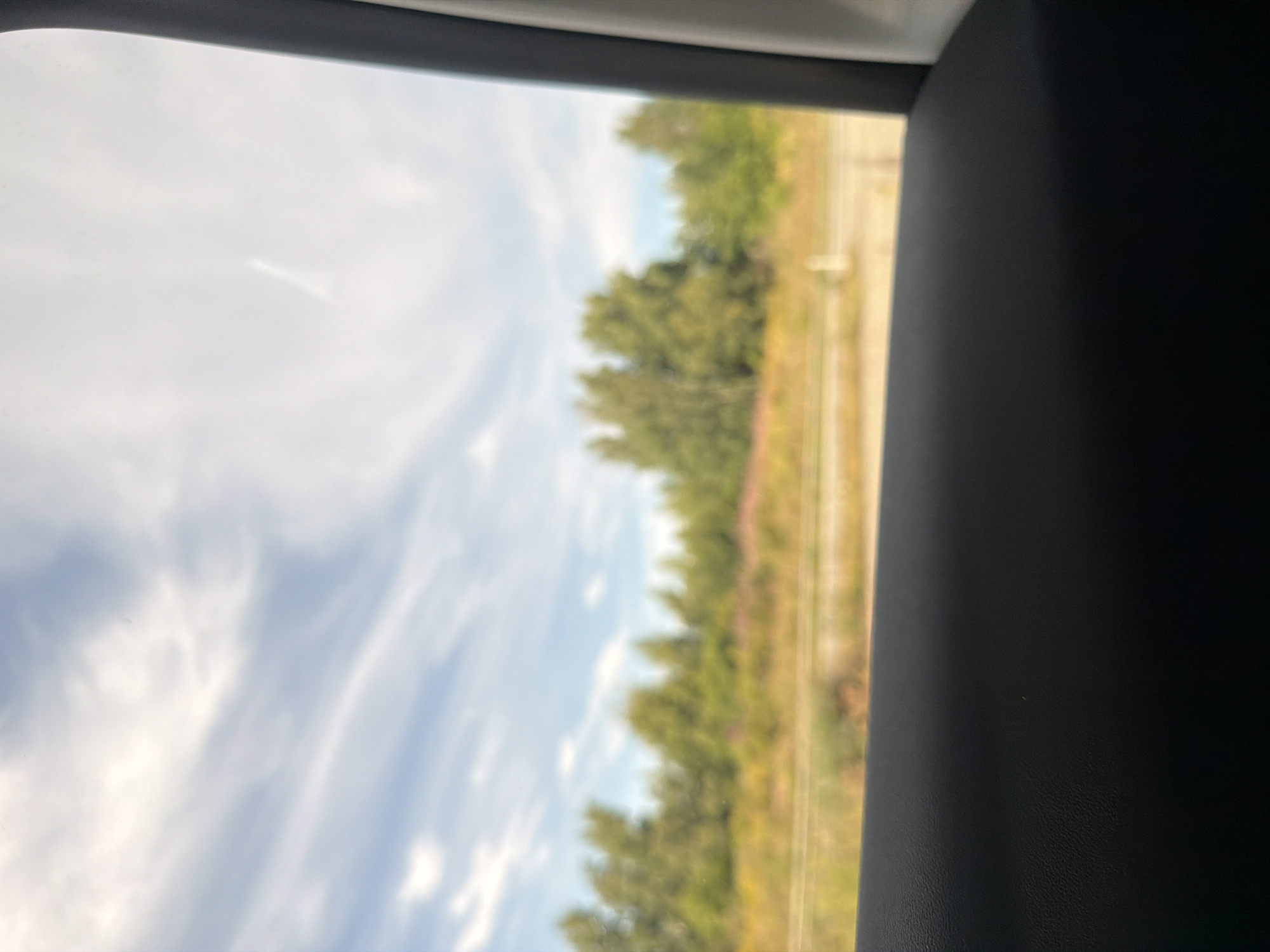 The image is an outdoor scene, viewed through what appears to be a car window. In the foreground, a dark, textured surface, likely the interior of a car door or armrest, occupies the bottom third of the frame. The middle ground shows a blurred landscape with trees and vegetation, suggesting a natural or semi-natural environment. Above the treeline, a blue sky with wispy white clouds is visible. The overall impression is of a journey through a rural or suburban area. The location is identified as Skumparp, Sweden. There are no people or specific actions depicted in the image.