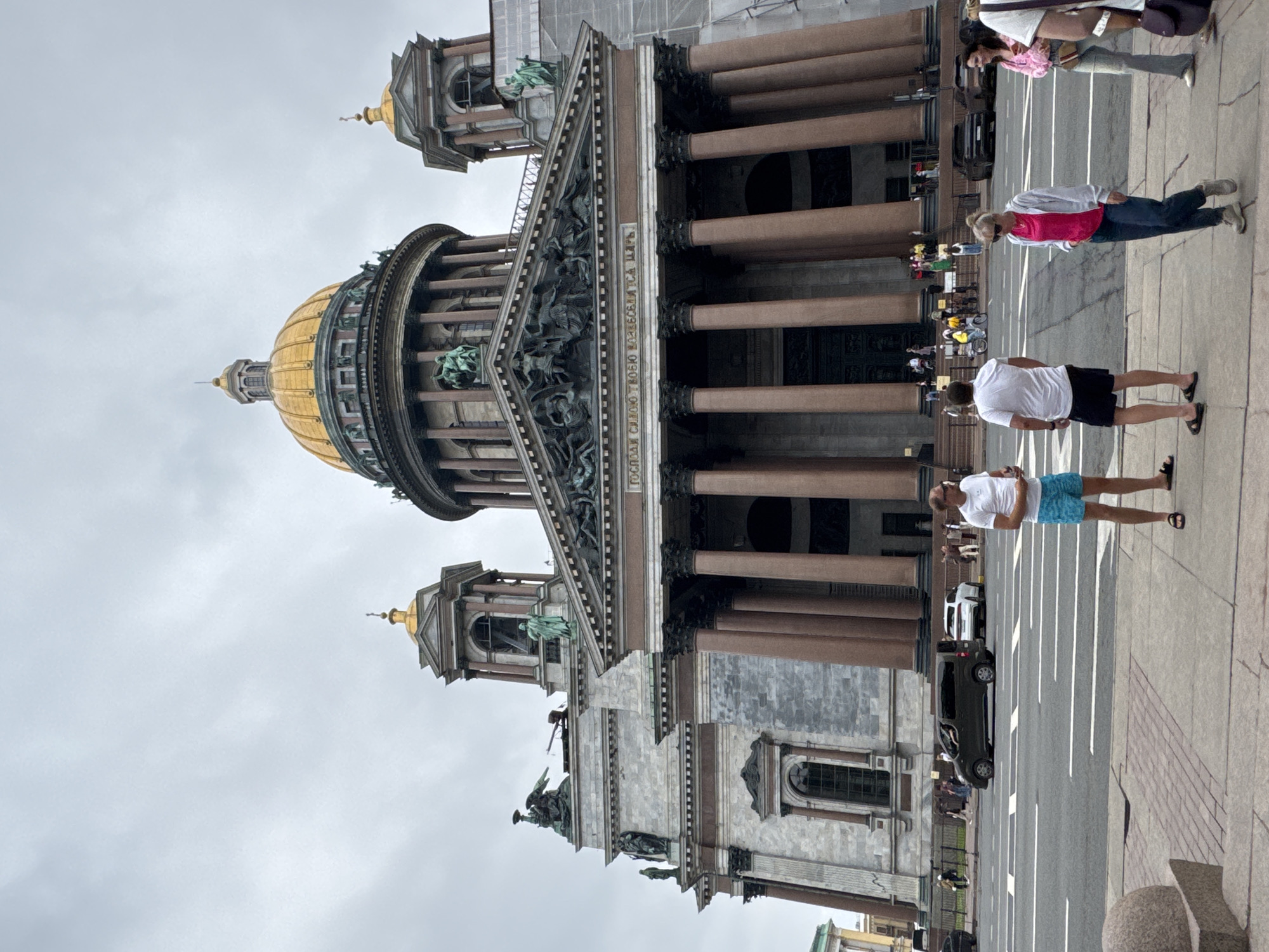 The image shows Saint Isaac's Cathedral in Saint Petersburg, Russia, under a cloudy sky. The massive neoclassical cathedral features a prominent golden dome and a grand portico with numerous columns. In the foreground, several people are standing on a sidewalk, facing the cathedral or walking. A street with passing cars is visible between the sidewalk and the cathedral. A dark green minivan is in the middle of the street, and a white SUV is behind it. Other people are seen in the background near the entrance of the cathedral, and some are on the street. Scaffolding is visible on the right side of the building, indicating ongoing construction or renovation.