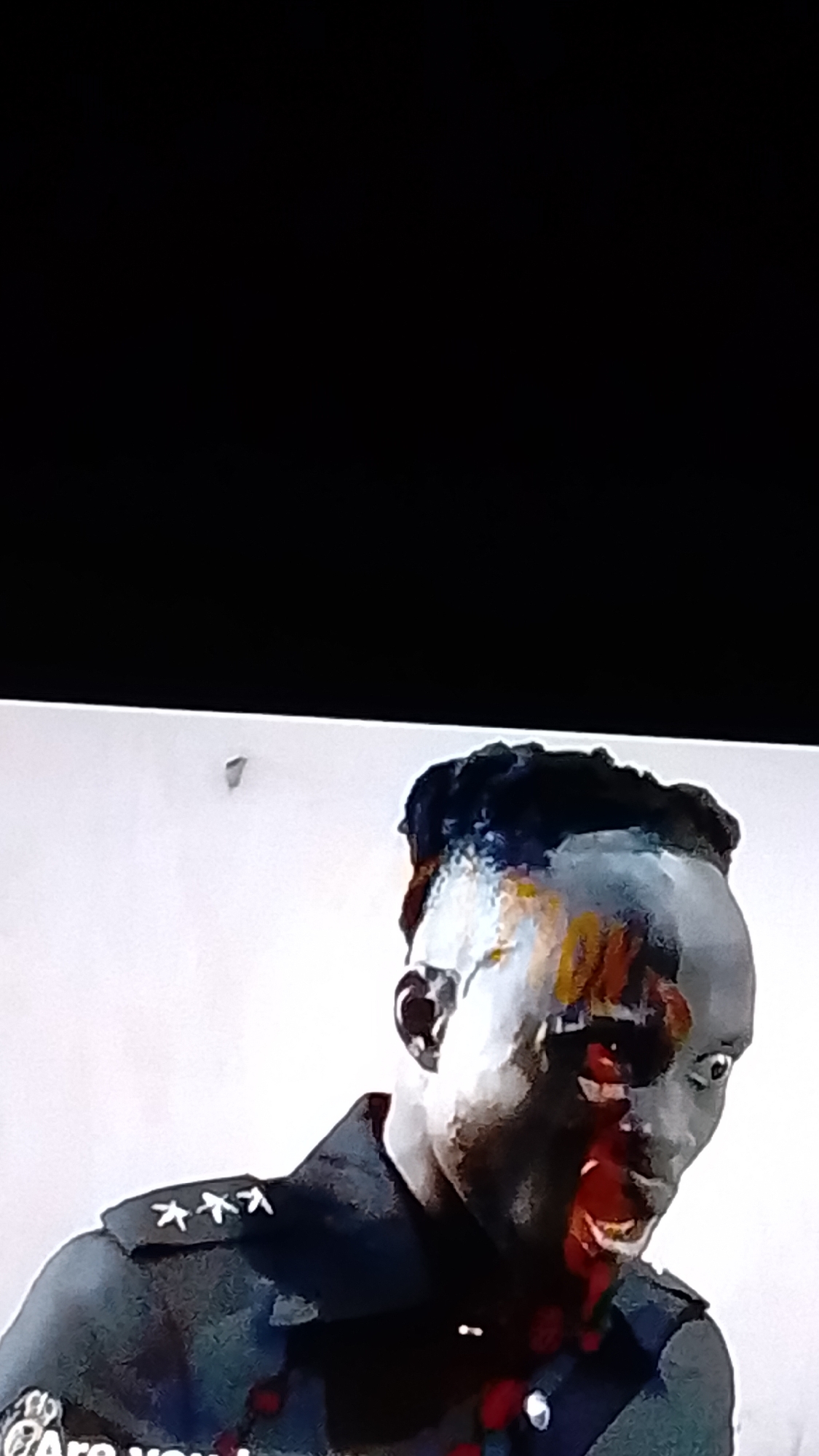 The image shows a close-up of a Black male wearing a dark uniform with silver insignias on the shoulder. His face is partially obscured by what appears to be blood or paint, with some splatters on his neck and uniform. His eyes are wide, and his mouth is open, suggesting distress or a reaction to something. The background is a plain, light-colored surface. The image is likely a still from a video or a heavily edited photograph, and there is no visible indication of its location in Lokoja, Nigeria, or any specific environmental context.