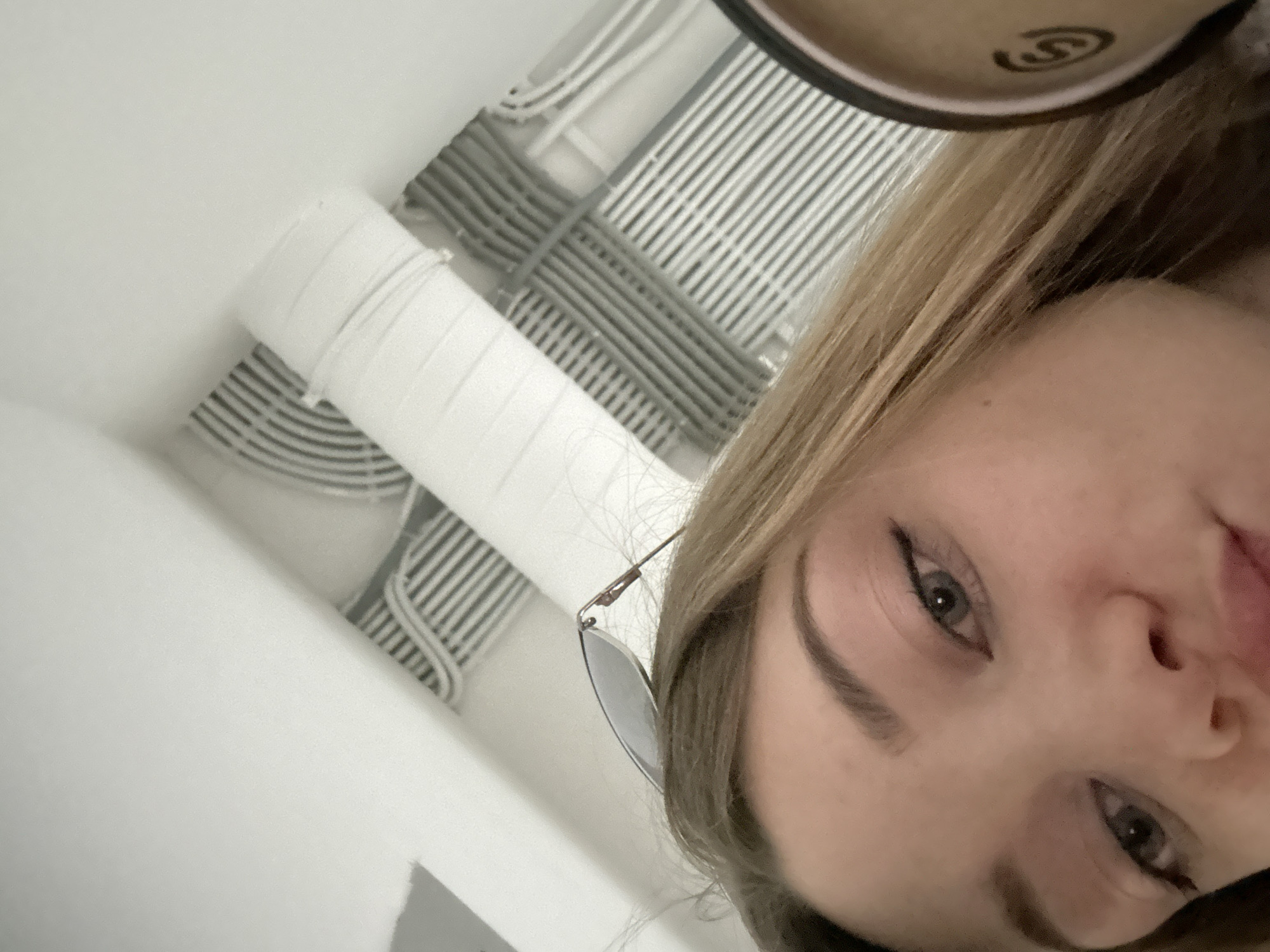 A close-up, tilted shot shows a fair-skinned person with blond hair, gray eyes, and sunglasses perched on the side of their face. They are indoors, with a white ceiling overhead featuring ventilation ducts and wrapped pipes. While the image gives no direct indication of Kazan, Russia, the location cannot be ruled out, as the scene itself is commonplace.