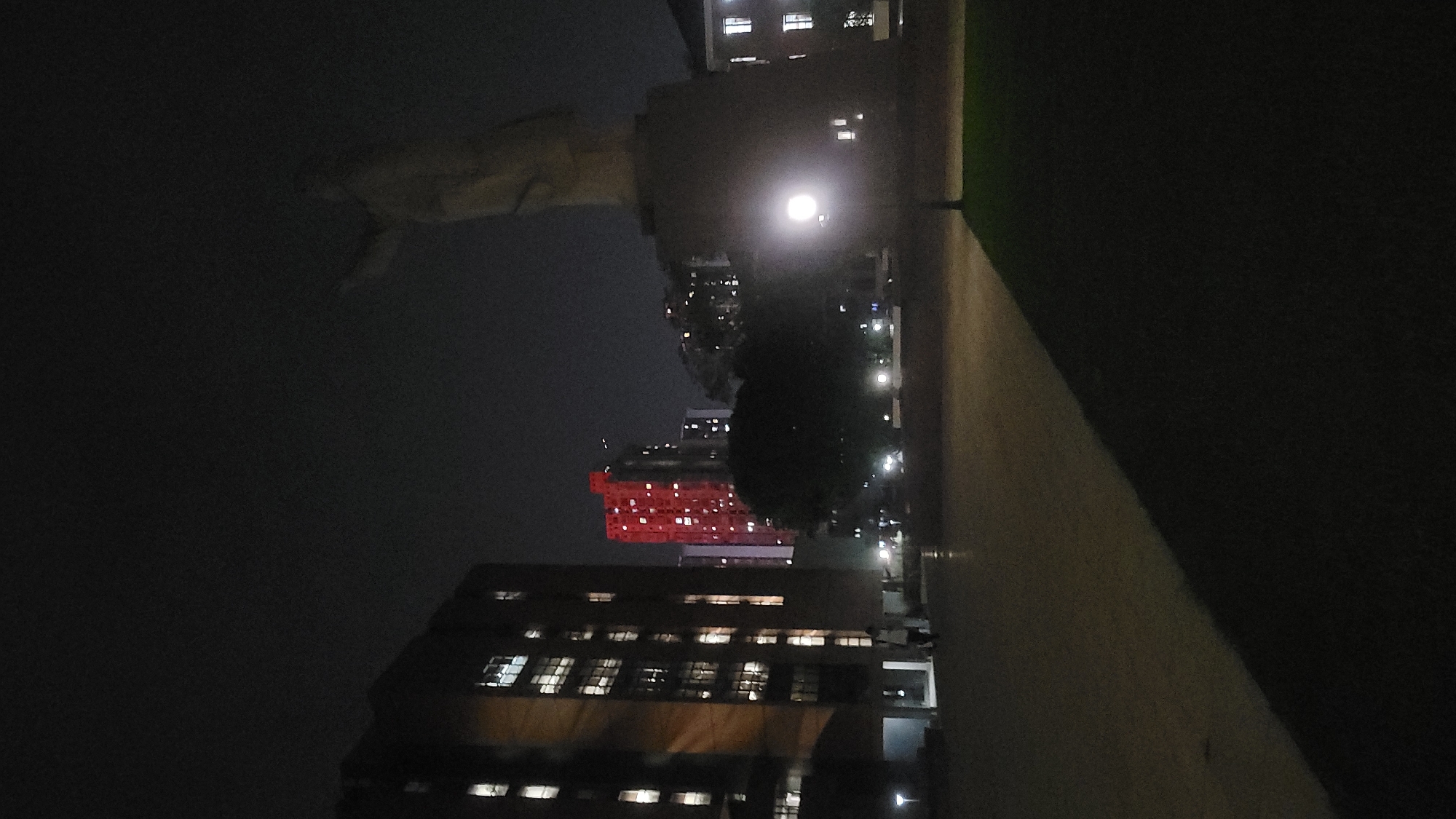 The image is a night shot of the urban landscape in Yuzhong District, China. Several buildings are visible, with some windows illuminated. One building stands out with its windows lit up in red. Streetlights are scattered throughout the scene, creating bright spots in the darkness. The sky appears overcast. The photograph is taken at ground level.