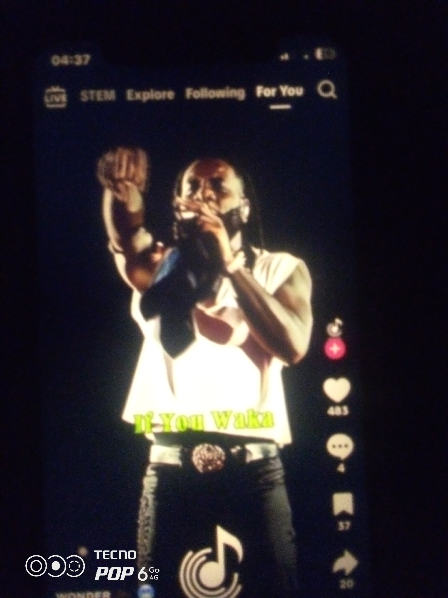 The image shows a man singing into a microphone, with his right fist raised. He is wearing a white sleeveless shirt and black pants with a decorated belt. Text overlay on the image reads "If You Waka" in a light green font. On the right side of the screen, there are icons representing likes, comments, shares, and saves, with corresponding numbers. At the bottom left, there is a logo that says "TECNO POP 6 Go 4G". The image appears to be a screenshot of a mobile phone screen, likely from a social media platform like TikTok. The top of the screen displays a time of "04:37" and navigation options such as "LIVE", "STEM", "Explore", "Following", and "For You", along with a search icon. The background is dark, suggesting a stage performance or a dimly lit environment. There is no specific information about the location in the city of Howayi, Nigeria, within the image itself.