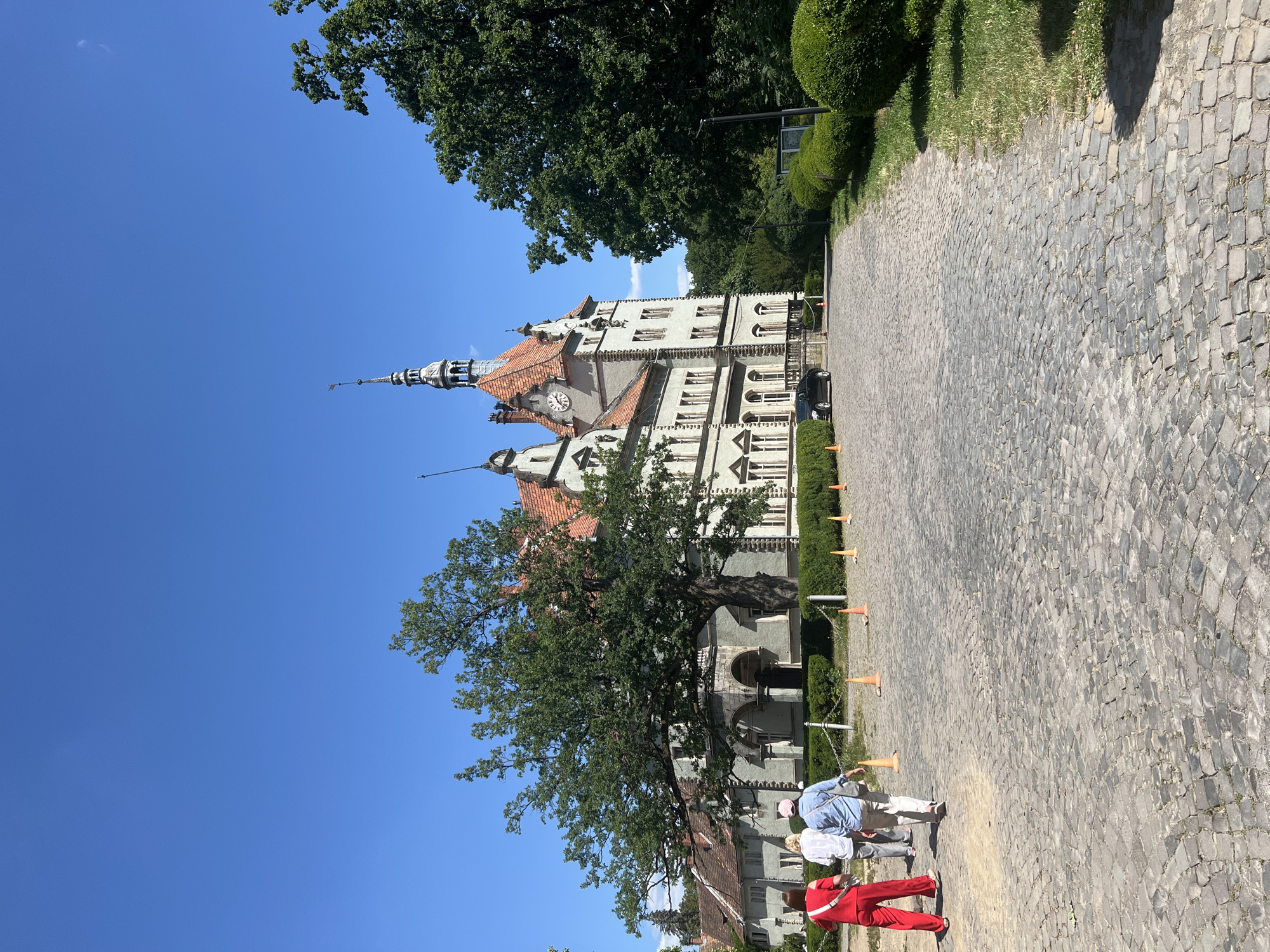 A paved, cobblestone pathway leads towards a large, stone building with a red-tiled roof and a prominent clock tower. Three individuals are walking away from the camera on the pathway, with one person in a red outfit and two others in lighter clothing. A large, leafy green tree stands to the left of the building. The sky is clear and blue. The scene appears to be set in a park-like environment with manicured bushes lining the right side of the pathway. The building has a historic architectural style.