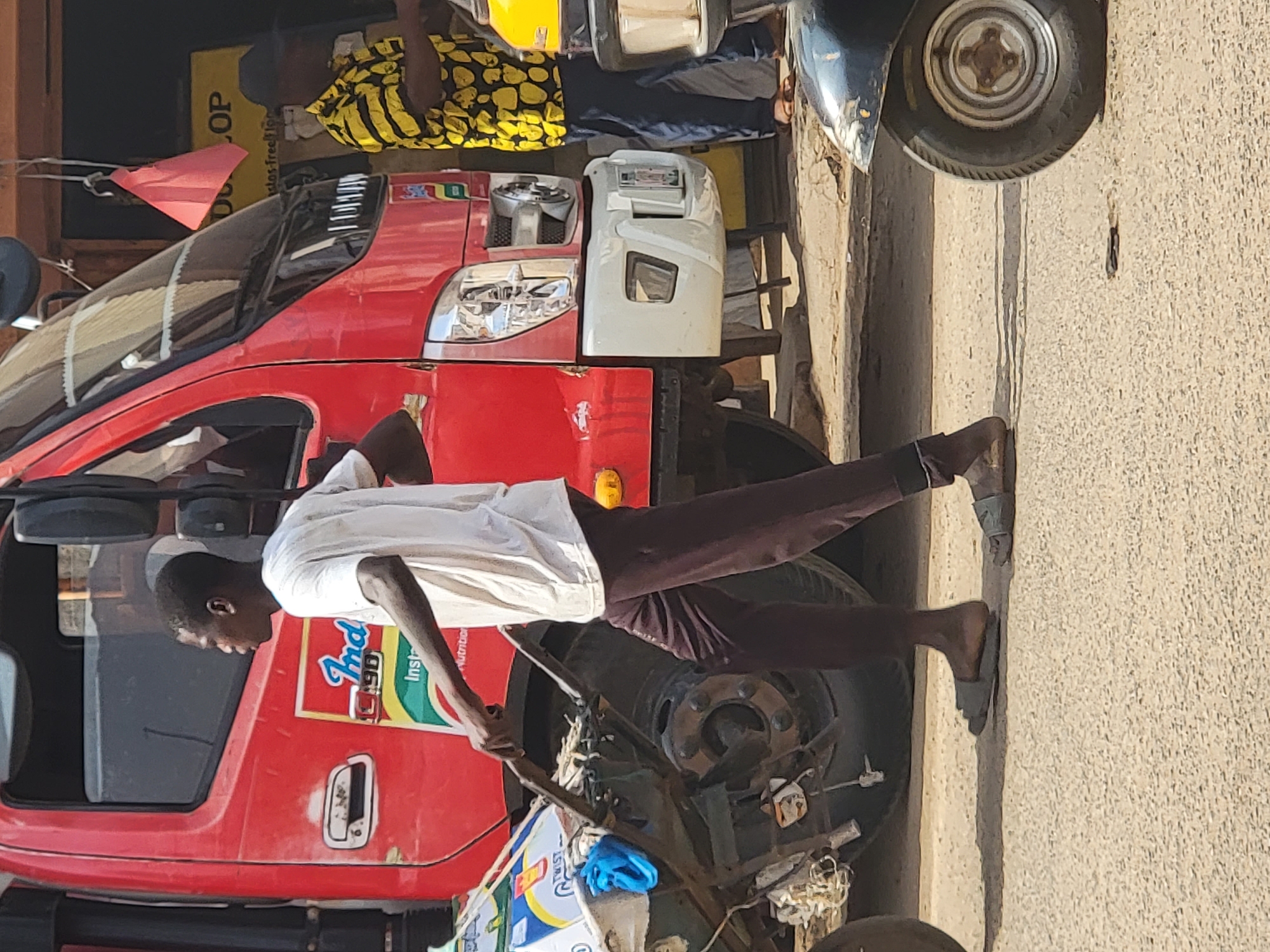 The scene shows a man in a white shirt and brown pants standing next to a red truck in Maiburiji, Nigeria. The truck has "Indomie Instant Noodles" branding on its side. Another man wearing a yellow patterned shirt and black pants is standing near the truck. Part of a motorcycle is visible, parked near the truck. The setting appears to be a street in Maiburiji.