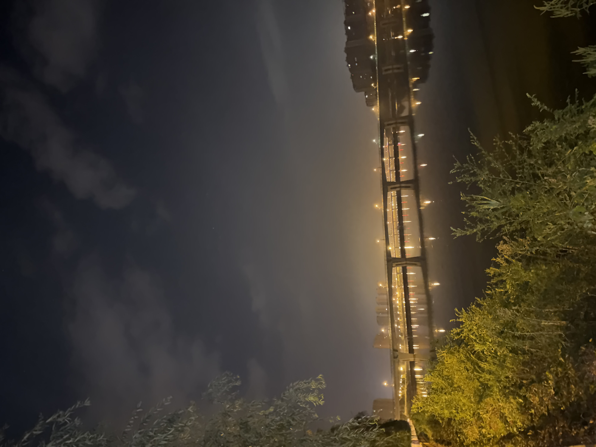A wide-angle, eye-level night shot captures a tranquil river scene in Taihe District, China. The dark sky is lightly clouded, with a few faint stars visible. A prominent multi-lane bridge, illuminated by numerous streetlights, stretches across the middle of the frame, its lights reflecting vibrantly on the calm water below. On the far bank, a dense cluster of apartment buildings, also lit up, forms a backdrop to the bridge. In the foreground, lush green vegetation lines the riverbank, with a weeping willow tree on the left casting dappled shadows. The overall atmosphere is peaceful and serene, with the glow of city lights contrasting with the natural elements.