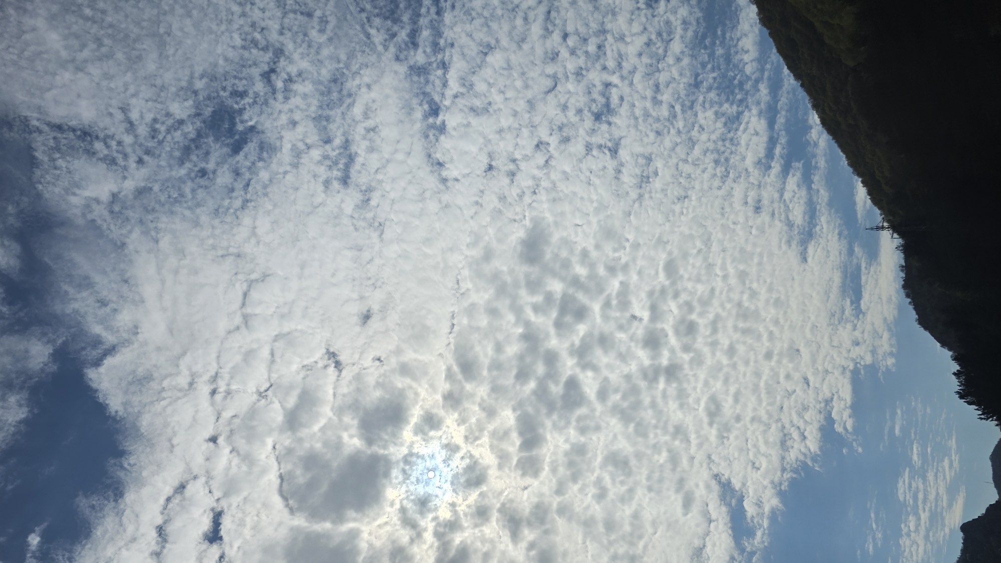 The image shows a sky filled with altocumulus clouds, with the sun visible through a gap. The right side of the image features a dark, silhouetted mountain ridge. The presence of a transmission tower cable supports the hypothesis that the location is Poleto, Bulgaria, known for its mountainous terrain and telecommunications infrastructure.