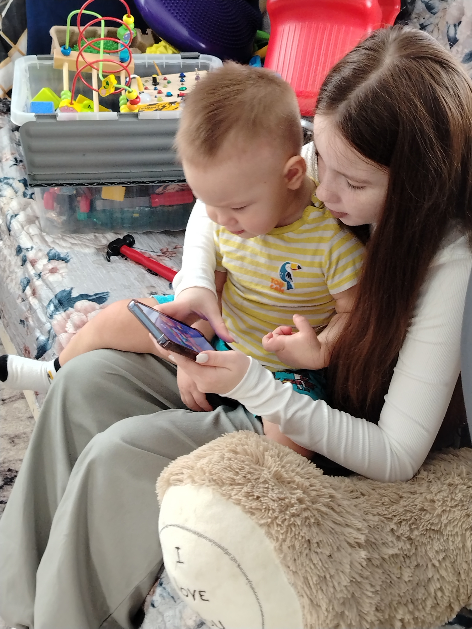 An indoor residential scene depicts two individuals interacting with a smartphone. A young female, estimated to be a child or teenager, with long brown hair, is seated wearing a white long-sleeved top and light green wide-leg pants. She holds a dark-colored smartphone in her left hand, displaying colorful content on its screen, and her nails are painted white. Seated on her lap is a male toddler with light brown hair, wearing a yellow and white striped t-shirt with a blue toucan graphic and blue patterned shorts, along with black and white socks. Both individuals are looking down intently at the smartphone screen.
In the background, on a patterned white and blue floral blanket covering what appears to be a bed or sofa, a clear plastic storage bin with a grey lid contains various colorful toys, including a bead maze and an activity board. A red toy hammer with a black head rests on the blanket nearby. A fluffy, light brown plush toy with "I OVE" embroidered on it is partially visible in the foreground. The overall environment suggests a casual, lived-in space within a residence. The location is an indoor residential setting in Unknown, Unknown.