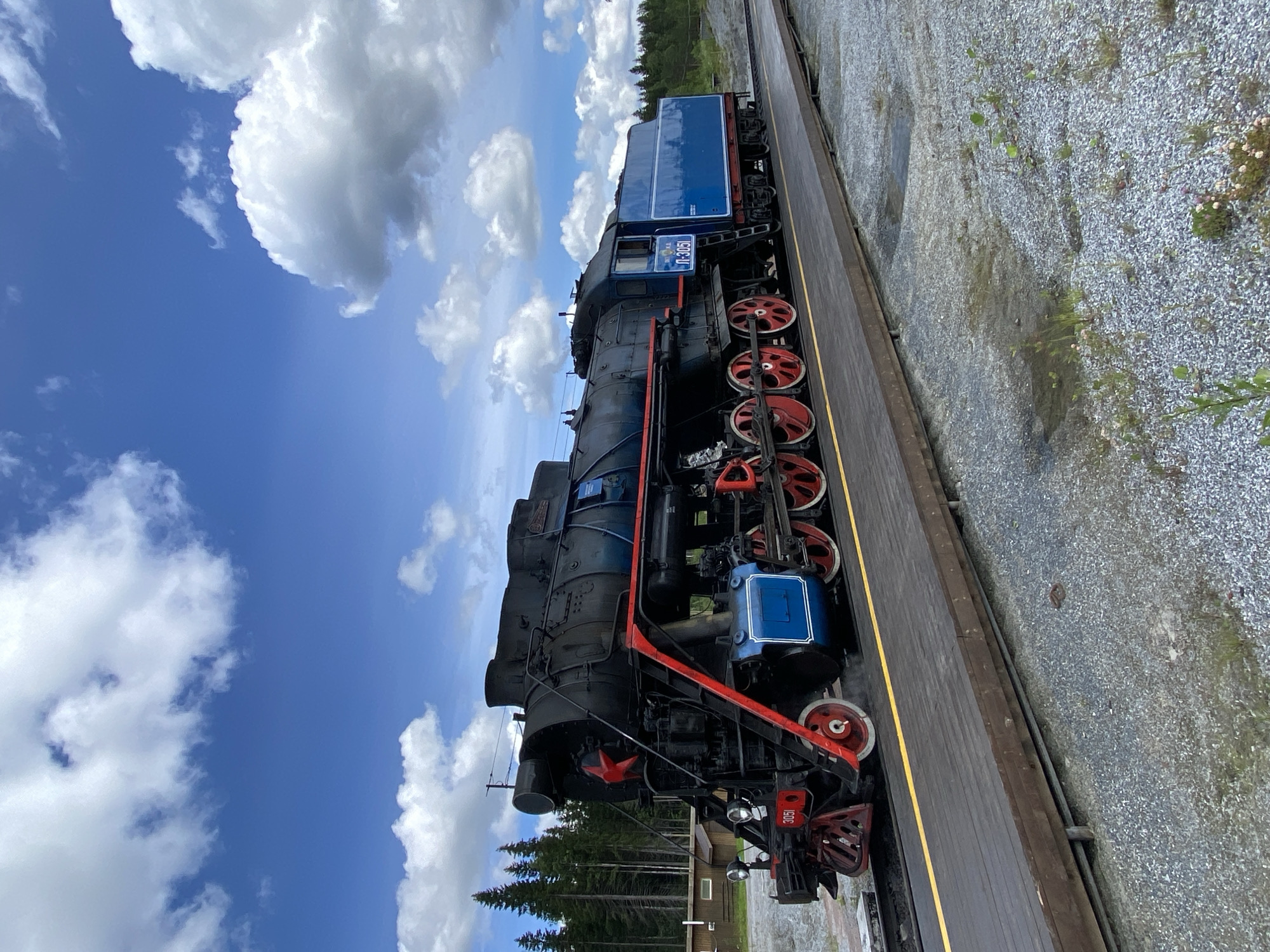 A vintage blue and black steam locomotive with red accents is parked on a wooden platform. The locomotive features a red star emblem on its front. The scene is set in the city of Ruskeala, Russia, with a forest of pine trees visible in the background. The sky is blue with scattered white clouds. The ground around the platform is covered in gravel and sparse vegetation. The image is a still photograph taken outdoors during the daytime.