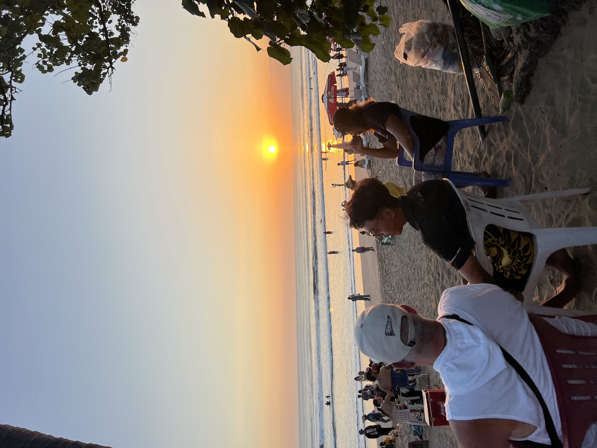 The image depicts a beach scene at sunset, likely in Kuta, Indonesia, given the context. The sky transitions from a pale blue at the top to a vibrant orange and yellow near the horizon, where the sun is setting over the ocean. Gentle waves are rolling onto the sandy beach.
In the foreground, three individuals are seated, facing the ocean. On the left, a person wearing a white sleeveless shirt and a white baseball cap is seated in a red chair. Next to them, in the center, is another person in a black t-shirt, seated in a white plastic chair. To the right, a third person, wearing a dark t-shirt and a baseball cap, sits in a blue plastic chair, holding a drink.
The beach is populated with other beachgoers, some of whom are in the water, while others are scattered along the sand. Beach chairs and umbrellas are visible further down the beach, suggesting a tourist area. Several objects are present on the sand in the foreground and midground, including a red cooler, a rake, a green bottle, and bags of trash. The overall atmosphere appears relaxed, with people enjoying the end of the day.