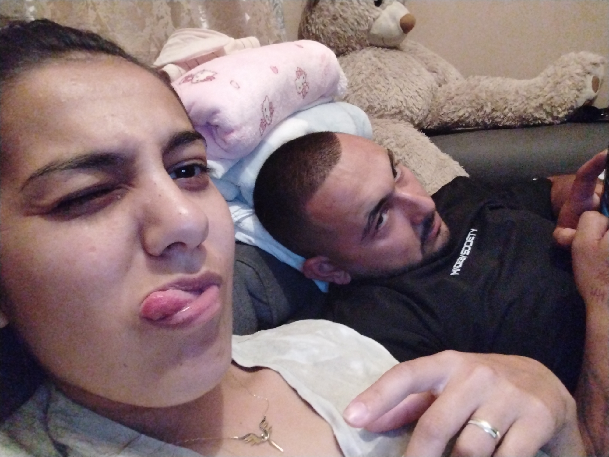A selfie shows a light-skinned woman winking with her tongue sticking out. A light-skinned man with closely cropped dark hair is reclining behind her and looking to the side, holding a dark object in his right hand. The man is wearing a black shirt with text that reads "MICRBY/SOCIETY" in white letters. He has a wedding ring on his left ring finger. In the background is a tan teddy bear, a pink patterned pillow, and a light-colored blanket. The location appears to be indoors, possibly on a couch, in an unknown city.