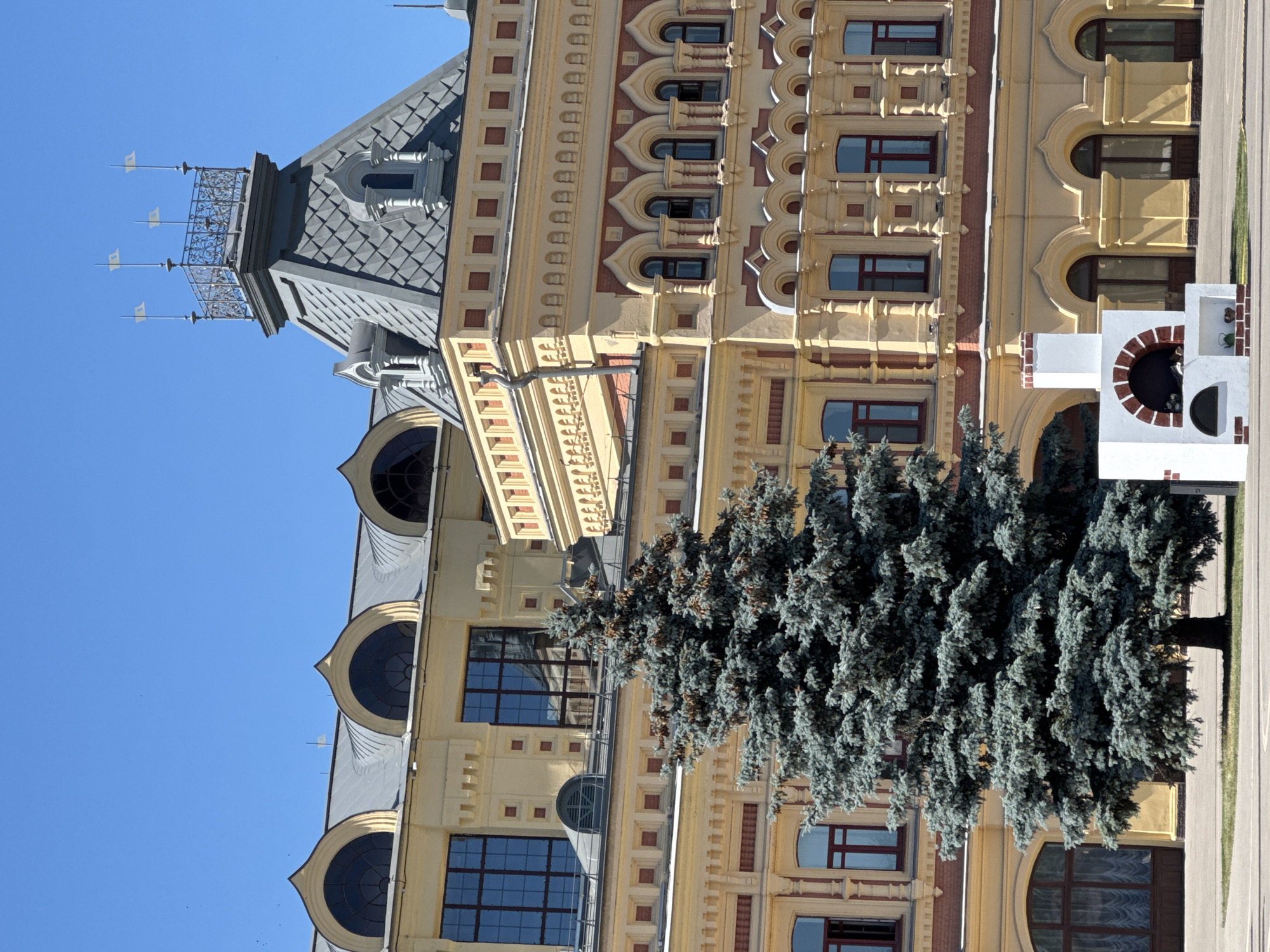 This is a photograph of a yellow and red ornate building with a grey, mansard-style roof and a decorative turret. A large blue spruce tree is situated in front of the building, obscuring a significant portion of its facade. The building features numerous windows, some arched and others rectangular, with dark panes. The architecture is detailed, with decorative moldings and recessed brickwork. On the right side of the building, a white structure with a red brick arched opening is visible. The sky is clear and blue, suggesting a daytime shot with good weather. The image is oriented vertically. The location is identified as Nizhny Novgorod, Russia.