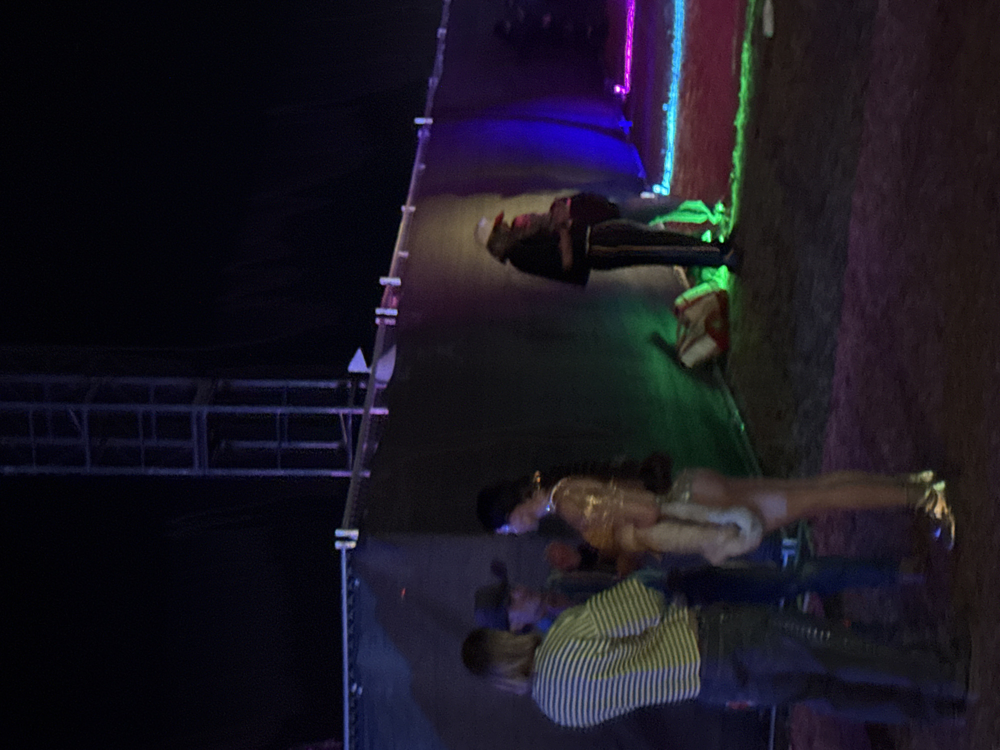 The image depicts a group of people near a stage structure in Thermal, California, United States. A male wearing a striped shirt and a female wearing a beige dress are standing and possibly talking. Another person in dark clothing is standing near the stage. The stage is illuminated with colored lights, including green, blue, and purple. There is also a metal support structure visible to the left. The ground appears to be dirt or gravel.