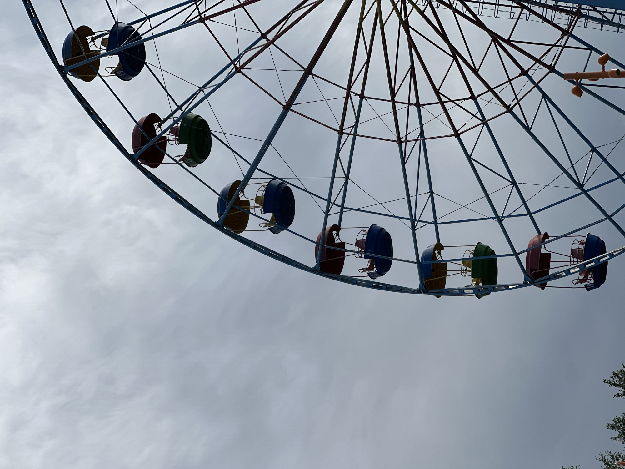 The image shows a Ferris wheel against a cloudy sky. The wheel is incomplete, with only a portion of it visible in the upper part of the frame. The carriages are painted in various colors, including blue, green, red, and yellow. The wheel's structure consists of a network of steel beams, some of which are blue and others red. The background is dominated by a gray, overcast sky. In the lower right corner, there is a hint of green foliage, possibly from a tree. The location is Unknown, Unknown.