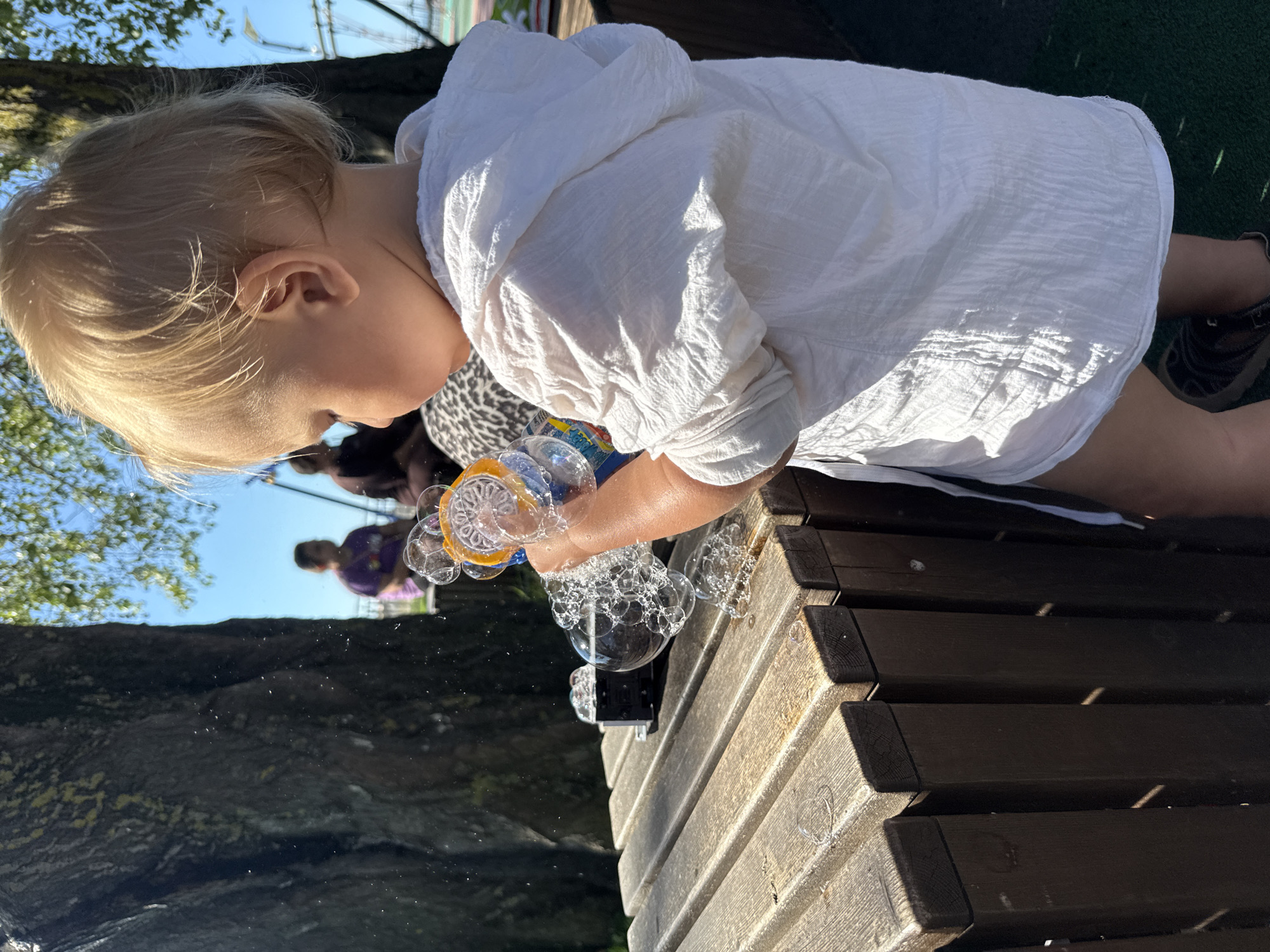 An infant with blond hair is playing with a bubble machine on a wooden bench. The infant is wearing a white hooded jacket and is holding the bubble machine with their right hand. Several bubbles are floating in the air around the infant and on the bench. In the background, two adults are sitting on a bench under a large tree. The sky is blue and there are some green leaves visible from the tree. The scene appears to be an outdoor recreational area, possibly a park or playground, located in the city of Sochi, Russia.