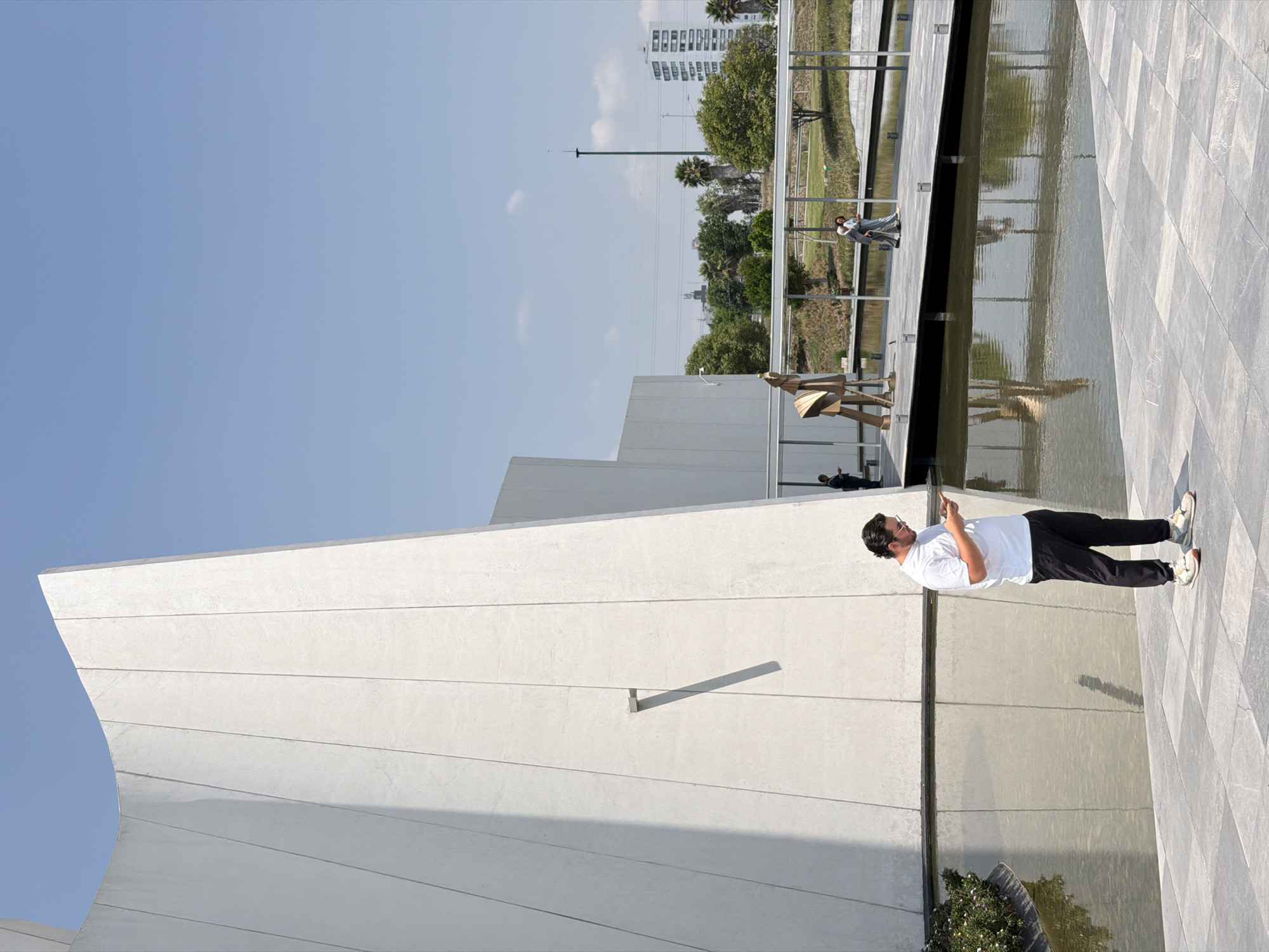 A medium shot captures a man in sunglasses and a white t-shirt, engrossed with his phone, standing to the side of a serene body of water. His presence complements the surrounding architecture, dominated by the Museo Internacional Barroco in San Bernardino Tlaxcalancingo, Puebla, Mexico. The museum's crisp white facade and unique, angled design are distinctly visible in the background. Other people are seen at a distance near the water. A row of trees stands in the background further away.