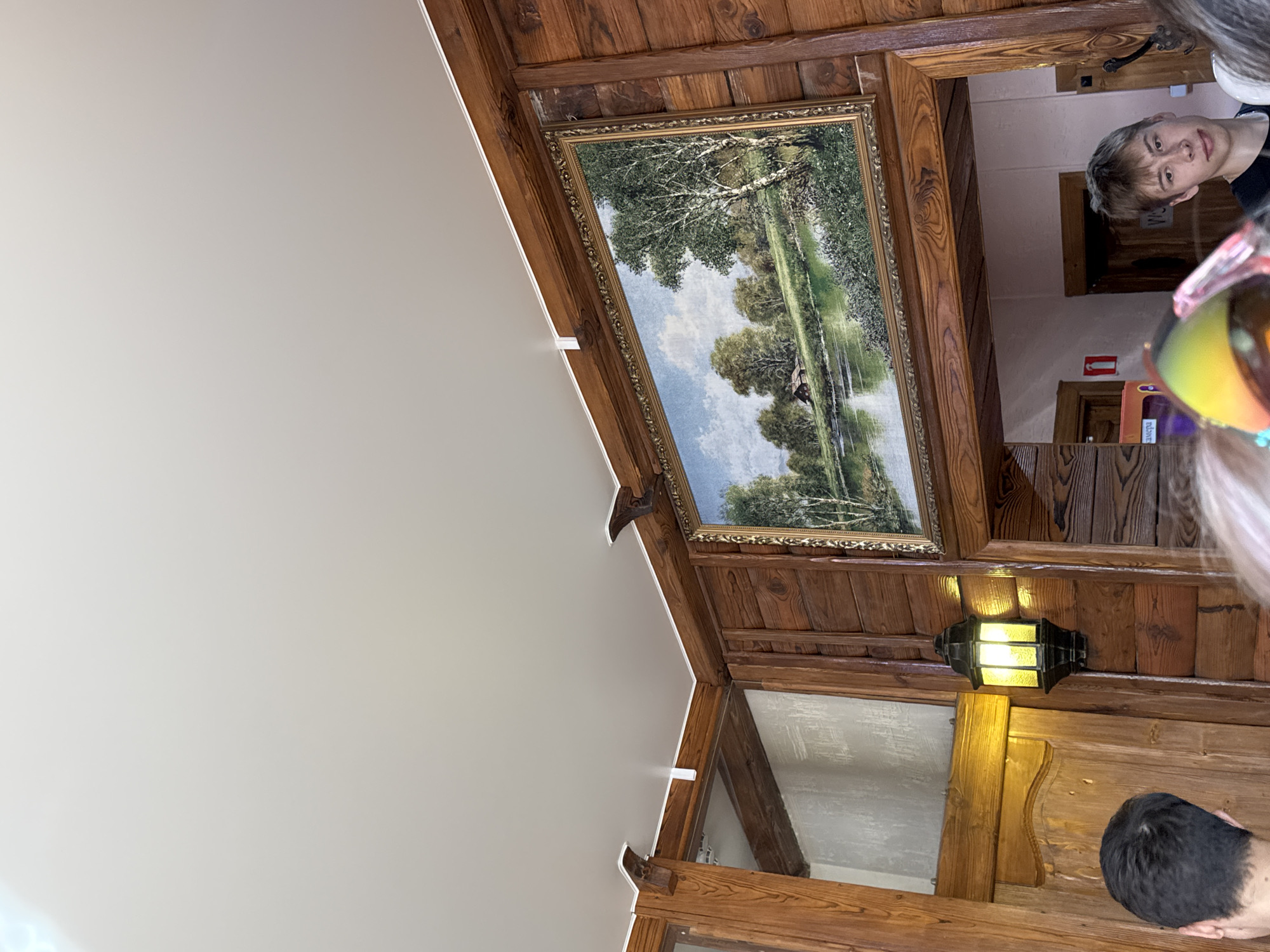 The image shows an interior space with wood paneling on the walls and ceiling. A framed landscape painting hangs on the wall. A lit lantern is attached to the wall below the painting. Two people are partially visible: one person is a young man looking at the camera, and another person is partially obscured. The location is likely Grishanki, Russia.