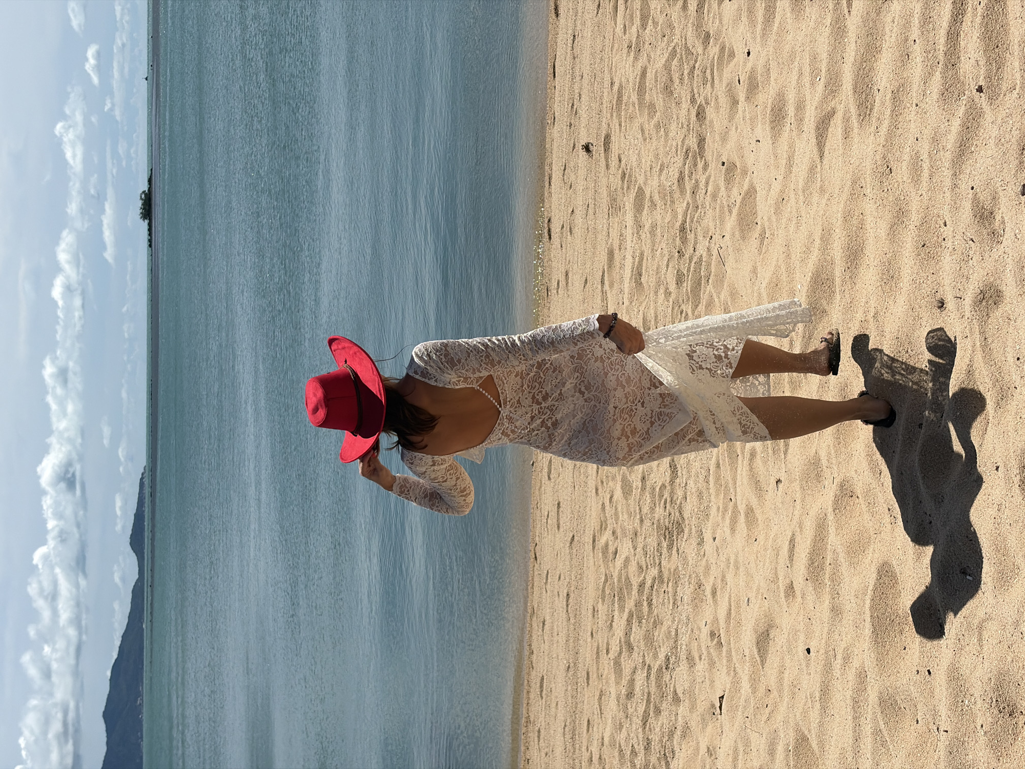 In Baan Bo Phut, Thailand, a woman stands on a sandy beach, facing away from the camera and towards the ocean. She is wearing a white lace dress with long sleeves and a low back adorned with pearls, and a bright red wide-brimmed hat. The ocean stretches out before her, a tranquil blue under a sky dotted with white clouds. In the distance, a chain of islands and hills form a hazy horizon line. The sand on the beach is light brown and textured with footprints and scattered debris. The woman's shadow is cast on the sand to her left.