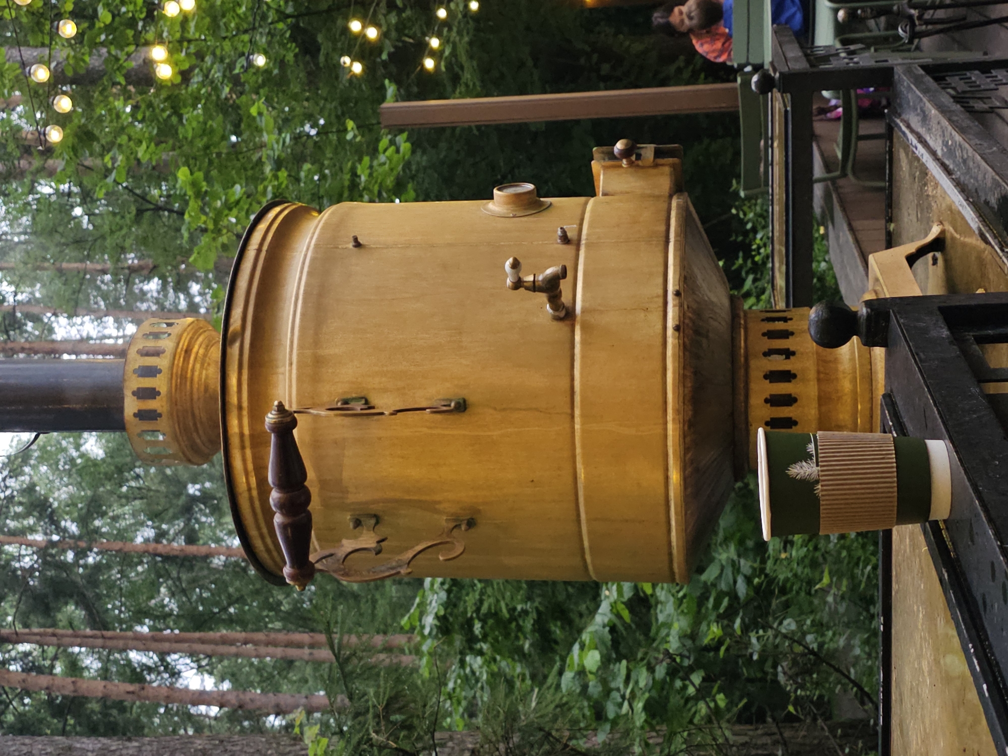 In Nemchinovo, Russia, a large, weathered, yellow samovar stands prominently in an outdoor setting with trees and string lights visible in the background. The samovar has decorative wooden handles and a spigot. In the foreground, a green disposable coffee cup with a ribbed brown sleeve sits on a wet surface. In the background, out of focus, two people, one adult and one child, are seated at a table. The overall environment suggests an outdoor cafe or recreational area.