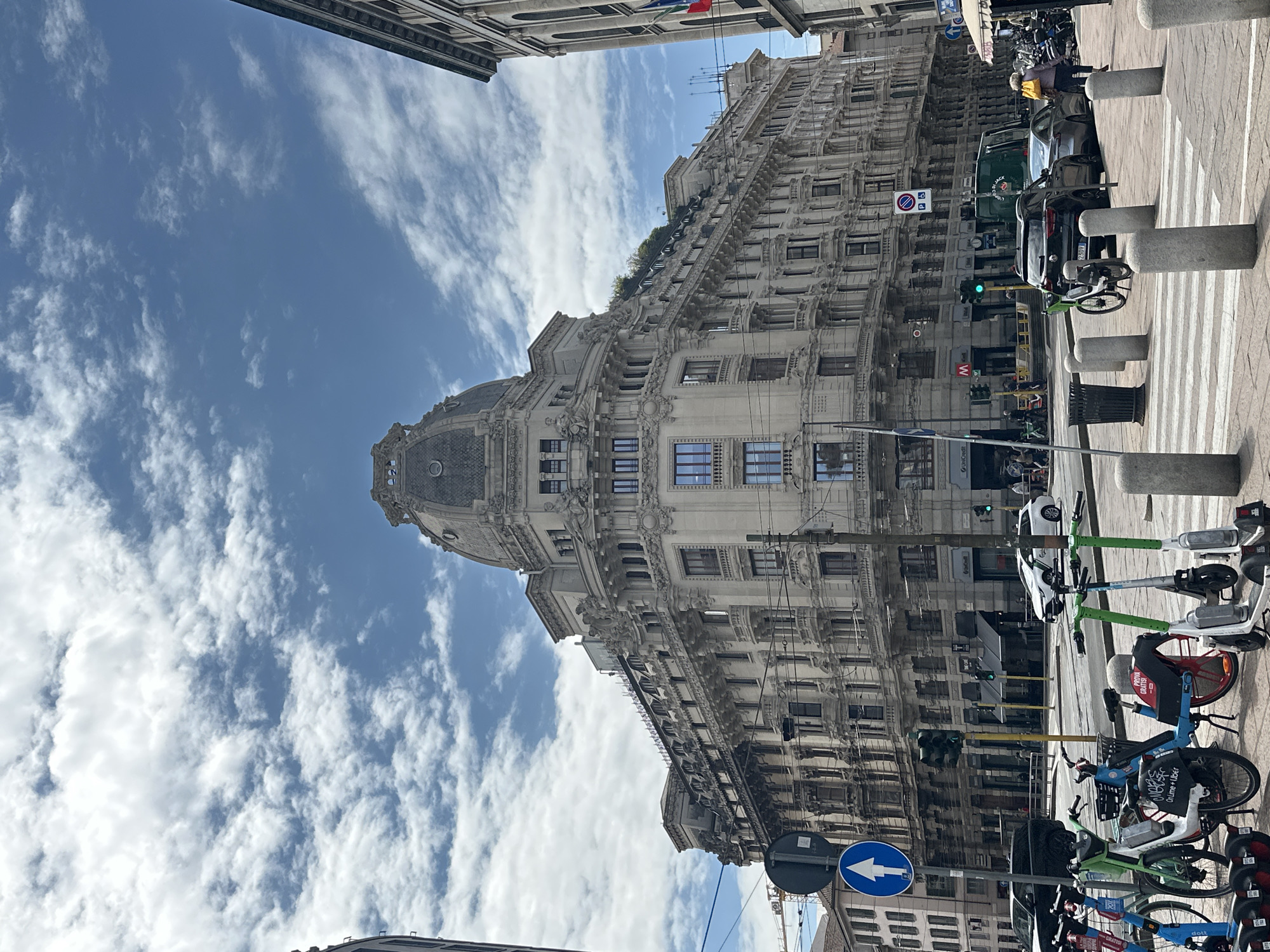 The image captures a vibrant street scene in Milan, Italy, under a bright blue sky with scattered white, wispy clouds, suggesting a clear and sunny day, likely mid-day.
The main subject is a grand, ornate building dominating the left side of the frame. It's a multi-story structure with intricate architectural details, numerous windows, and a distinctive domed corner at the front. The building appears to be made of light-colored stone or stucco, and some greenery is visible on one of its upper terraces.
On the right side, a bustling urban street or pedestrian area is visible, featuring a crosswalk marked with white stripes and several concrete bollards. A variety of vehicles are present, including parked cars (a dark sedan with license plate "239BV" and a green van), and a dense collection of shared mobility devices. These include numerous electric scooters in various colors (green, white, red, black) with brand names like "LIME", "TIER", and "DOTT", as well as shared bicycles (blue and red) with branding such as "PROVA GRATIS!" and "ROLLME + UBER". One person wearing a yellow jacket is partially visible on the sidewalk amidst the parked vehicles.
Other notable details include traffic lights, a blue circular street sign with a white arrow pointing left (when the image is viewed upright), a red and white "no parking" sign, and an "M" sign indicating a Metro station. Partially visible text includes "Unicredit" on a sign in the lower left corner. Overhead wires crisscross the upper part of the scene. The overall activity depicts typical urban life with transportation and pedestrian movement.