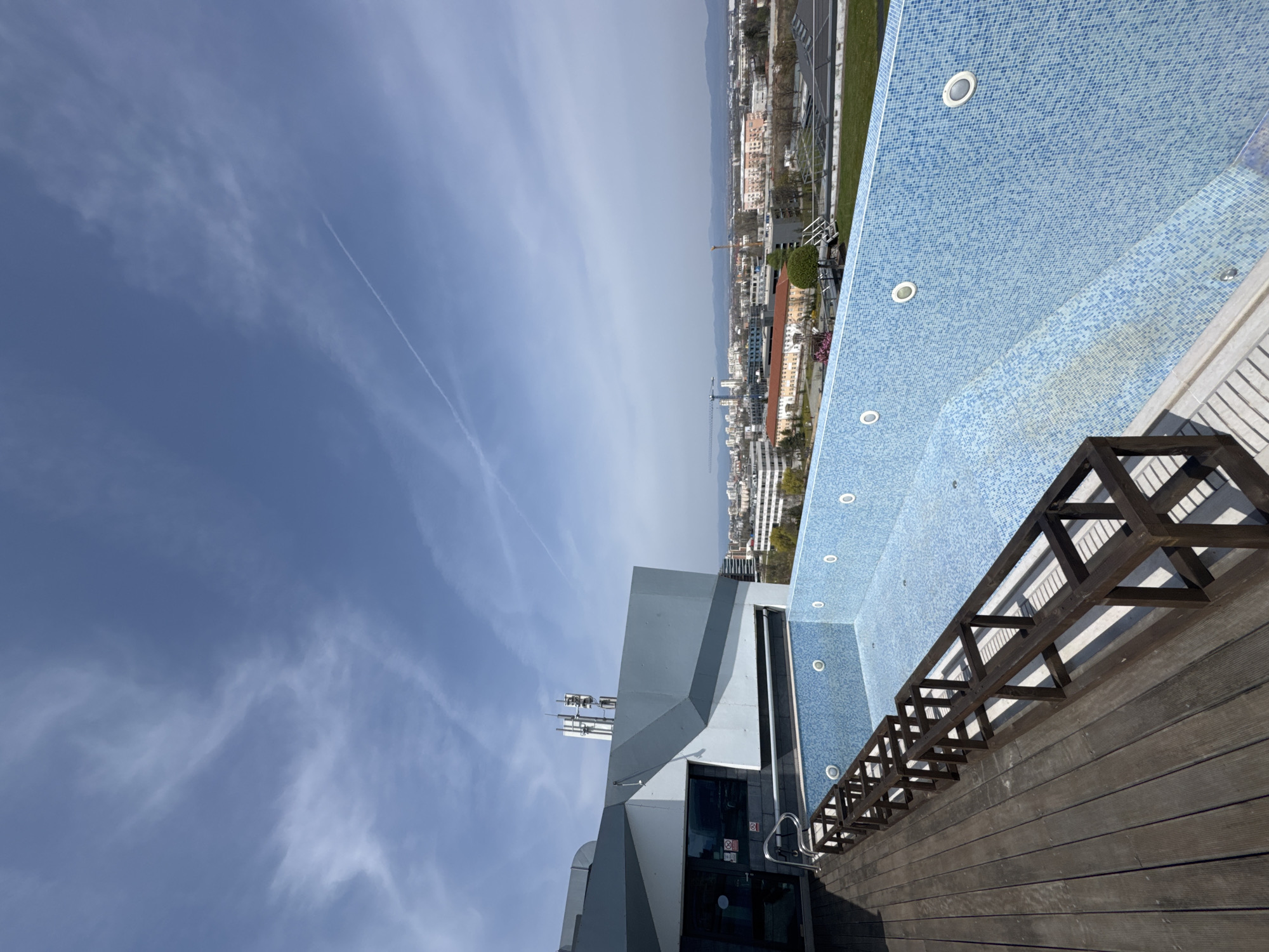 The image depicts a rooftop scene in Sofia, Bulgaria. A long, narrow swimming pool lined with small blue tiles runs alongside a wooden deck area with a dark wooden railing. The pool has several recessed lights along its side.
Next to the pool area, a modern building with a gray and white geometric facade is visible. The building appears to have telecommunications equipment installed on its roof.
In the background, the cityscape of Sofia stretches towards the horizon, with various buildings visible under a partly cloudy sky. A mountain range is also discernible in the far distance.
