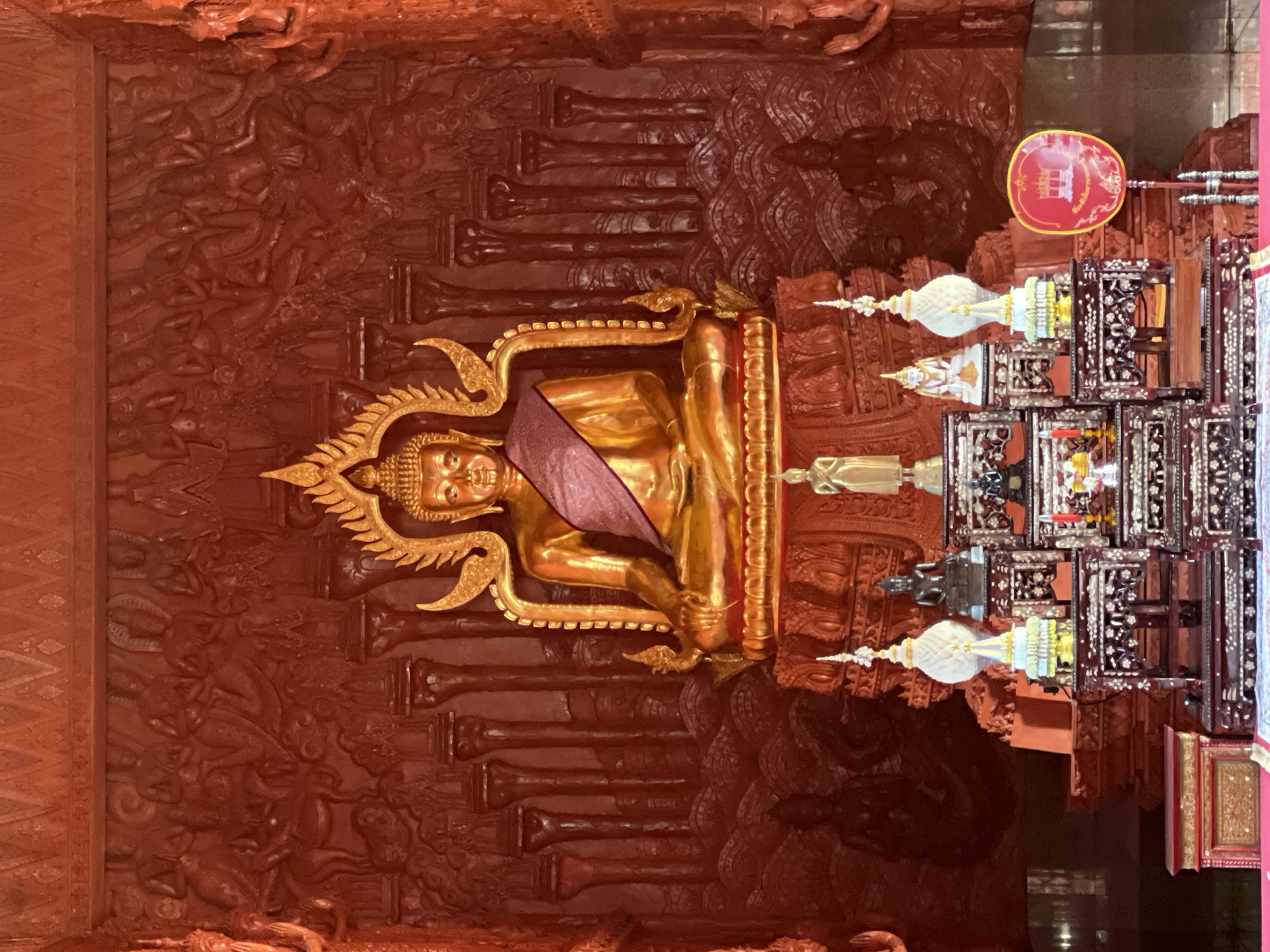 This image depicts the interior of a temple in Baan Hua Thanon, Thailand. The central focus is a large, golden Buddha statue seated on a tiered, ornate pedestal. The Buddha is adorned with a purple sash draped across its shoulder and chest. The background is a richly carved, red-toned wall featuring intricate reliefs of figures and architectural elements, possibly depicting scenes from Buddhist mythology or history.
In front of the main Buddha statue, a smaller altar is set up with various offerings and decorative items. These include smaller Buddha statues, tiered ornamental structures, and what appear to be ceremonial objects. To the right of the altar, a red circular fan with a central emblem is displayed. The overall color palette is dominated by warm reds and gold, creating a sacred and opulent atmosphere. The lighting appears to be ambient, highlighting the textures and details of the temple's decor.