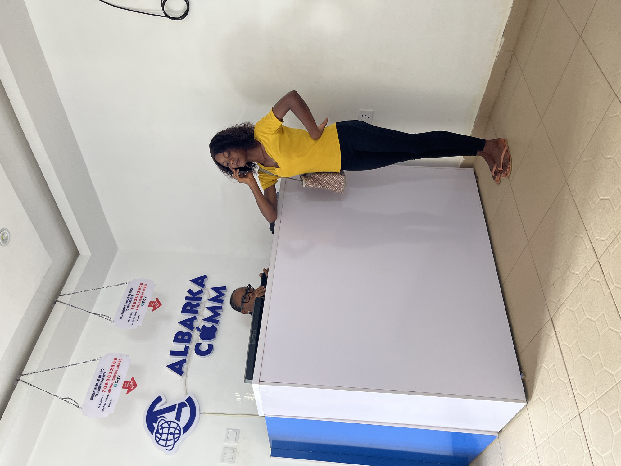The image depicts the interior of an Albarka Comm office. A young Black woman in a yellow T-shirt and black leggings is standing behind the reception counter, talking on her mobile phone. She has a purse slung over her shoulder. A second person, wearing glasses, is partially visible behind the counter.
Signage indicating payment instructions for Albarka Comm, with the account name and number (AUWAL ISHEHU AHMED, 7063632909), hangs from the ceiling on the left. The walls are white. The floor is tiled with a light-colored geometric pattern.