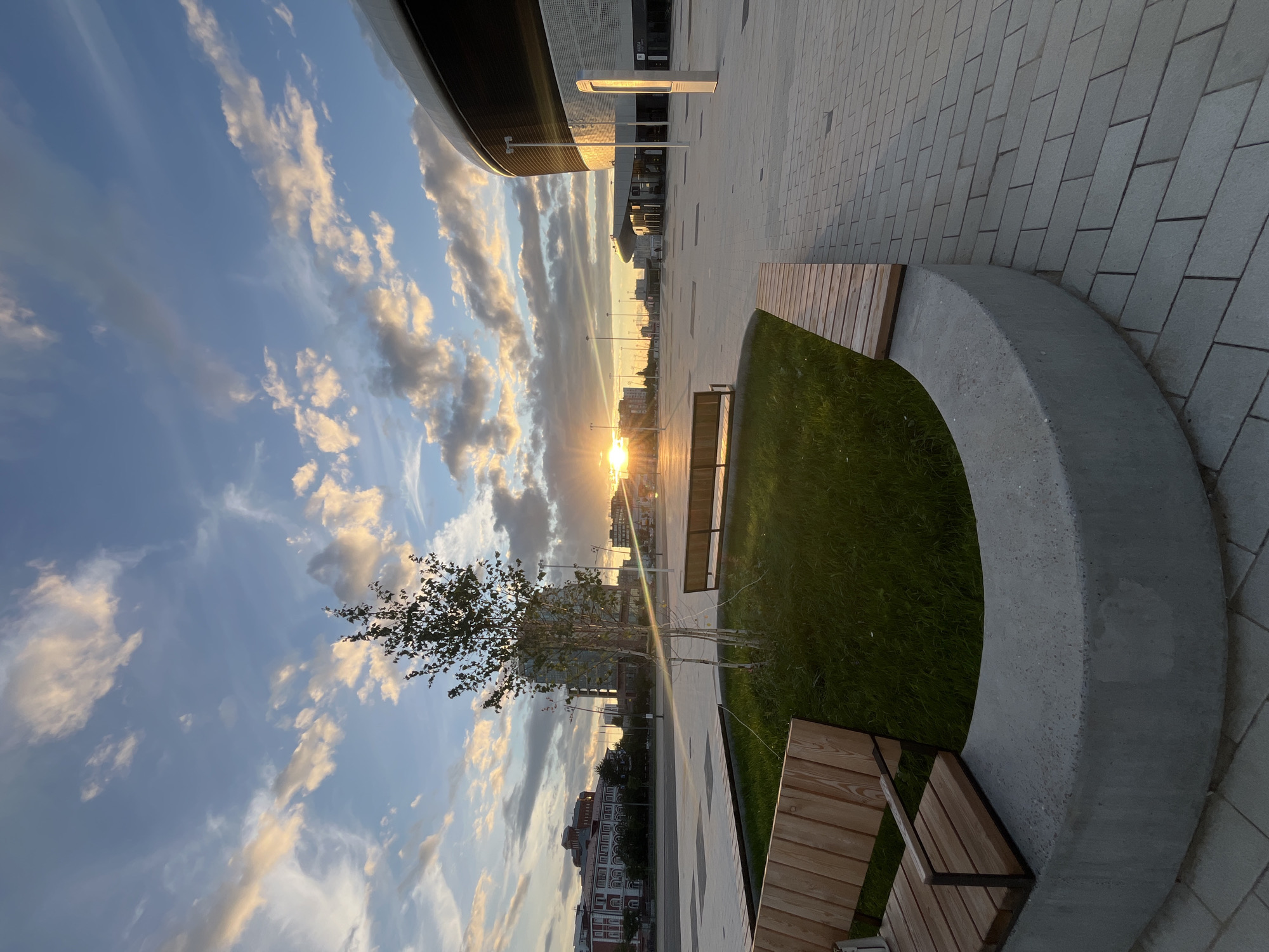 An outdoor, ground-level, wide-angle, eye-level shot captures a modern urban plaza at sunset. The sun, a bright orb just above the horizon, casts a warm glow across the scene, illuminating scattered clouds with hues of orange and pink against a blue sky. The foreground features a curved concrete planter with a patch of vibrant green grass and a young tree with thin branches. Two wooden benches with metal frames are positioned within the planter area, one in the lower left and another further back in the center.
The plaza itself is paved with light gray rectangular bricks laid in a herringbone pattern. To the right, a large, modern building with a curved, metallic facade dominates the background, its sleek design contrasting with the softer light of the sunset. In the distance, a row of multi-story buildings, some with red brick facades, line the street, suggesting a cityscape. Streetlights are visible throughout the plaza. The overall impression is one of a tranquil, contemporary public space bathed in the soft light of dusk.
This location is the Nizhny Novgorod Stadium (Nizhny Novgorod Sports Palace), located in Nizhny Novgorod, Russia.