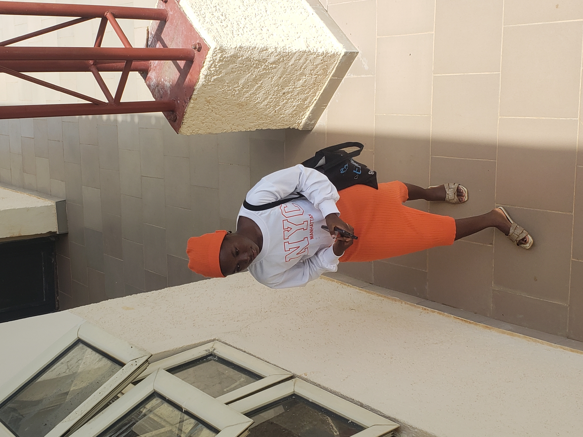 An overhead shot captures a young person wearing an orange beanie and a white long-sleeved shirt with "NYC MANHATTAN" written on it in red. They are also wearing an orange ribbed skirt and gold sandals. A black bag is slung over their left shoulder. The individual is standing on a paved walkway with large, light-colored square tiles, casting a shadow from a red metal structure. To the left, a textured, cream-colored wall features a window with multiple panes. The background shows more tiled flooring and a dark doorway. This scene is located in the city of Gumel, Nigeria.