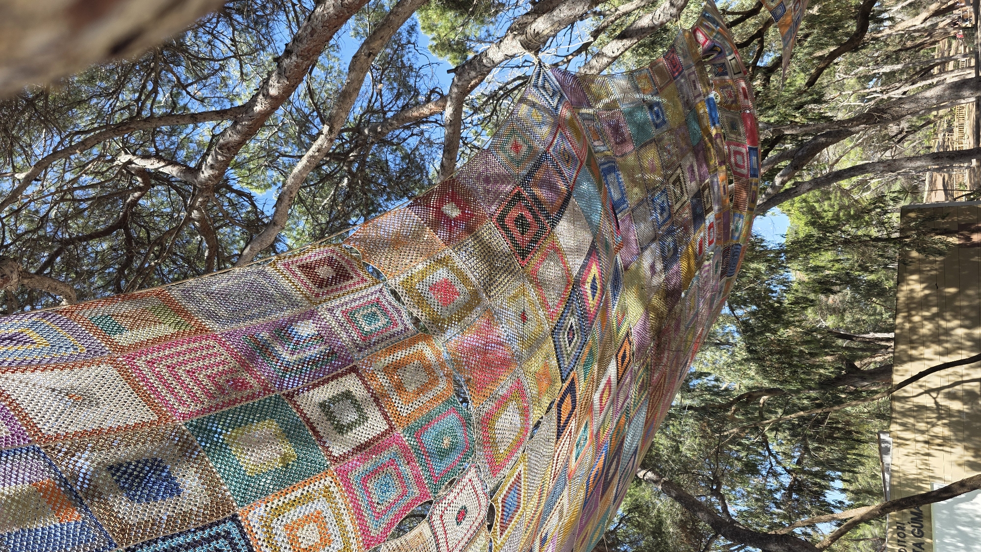 A low-angle, full shot shows a large, crocheted blanket hanging between tree branches under a sunny, blue sky. The blanket is made of numerous square, diamond patterned crocheted panels of various colors, including red, pink, yellow, green, blue, and brown. The blanket appears to be draped or suspended in a natural environment. The tree branches are mostly bare, though with some green foliage visible. In the background to the right, there is a beige building with the inscription "Auditori". The image was taken in Guardamar del Segura, Spain.