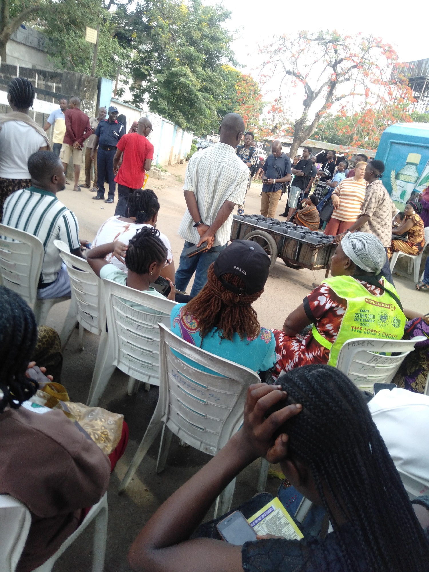 The image shows a gathering of people in Lagos, Nigeria. Many individuals are seated on white plastic chairs, while others stand in groups. A man pushes a small cart loaded with rectangular objects covered in black plastic. A police officer is visible in the background. A tree with bright red flowers is present in the scene. A sign on a woman's vest advertises a December 25th and 26th, 2025, event at 8 am, hosted by "The Lord's Chosen." A "PRADA" branded baseball cap is visible. There is a blue van with the word "POKAZ" visible.
