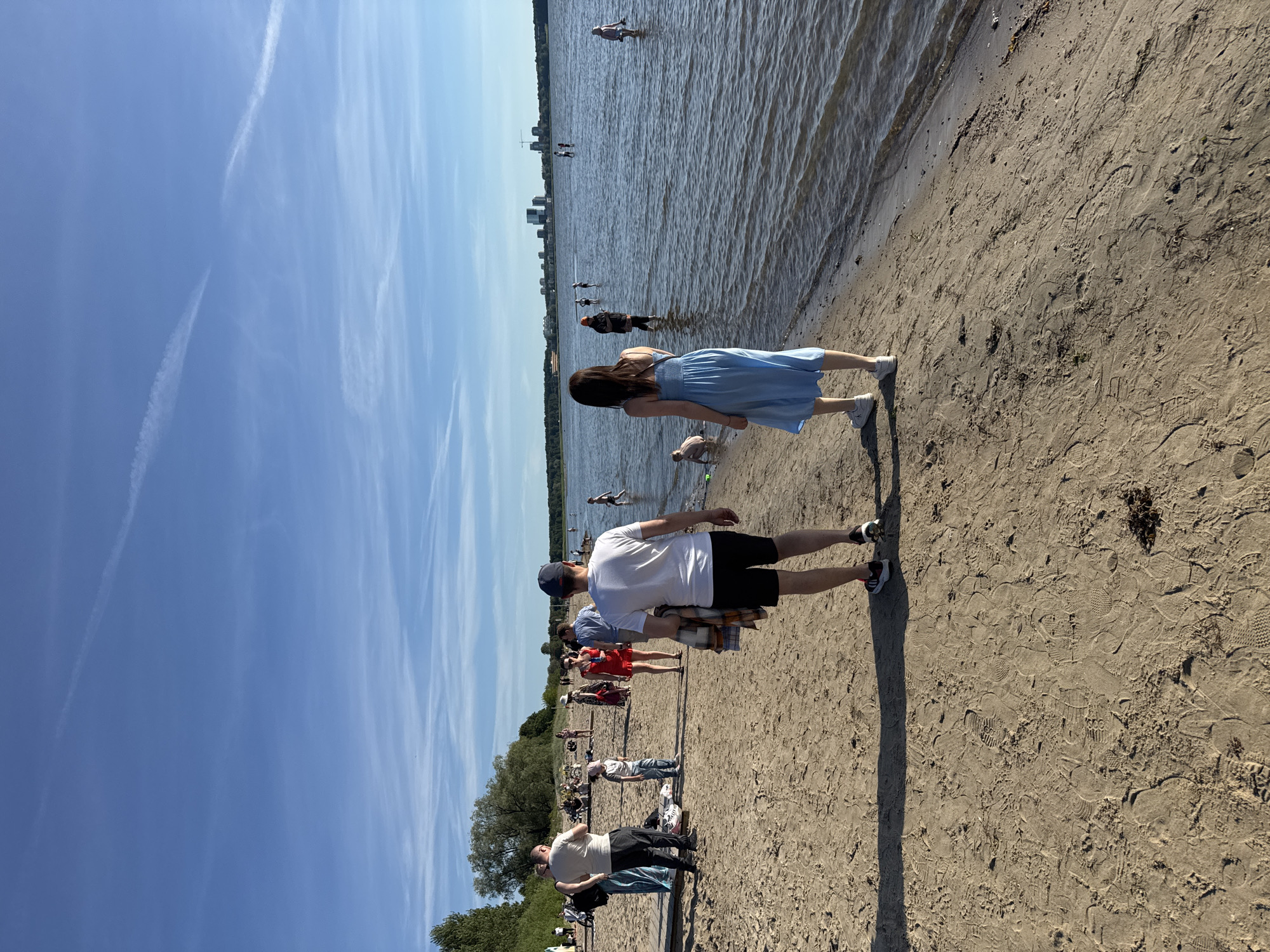 The image shows a sunny day at a beach in Tallinn, Estonia, likely Pirita Beach given the distant city skyline. The foreground features a sandy beach with numerous footprints. Two individuals are walking away from the camera on the beach, a man in a white t-shirt and black shorts, and a woman in a light blue dress. In the background, other people are scattered on the beach and in the water. Some are wading, while others appear to be swimming or playing. The water is calm, reflecting the blue sky with some wispy clouds. The city skyline, with its modern buildings, is visible in the distance across the water. Trees line the far shore.