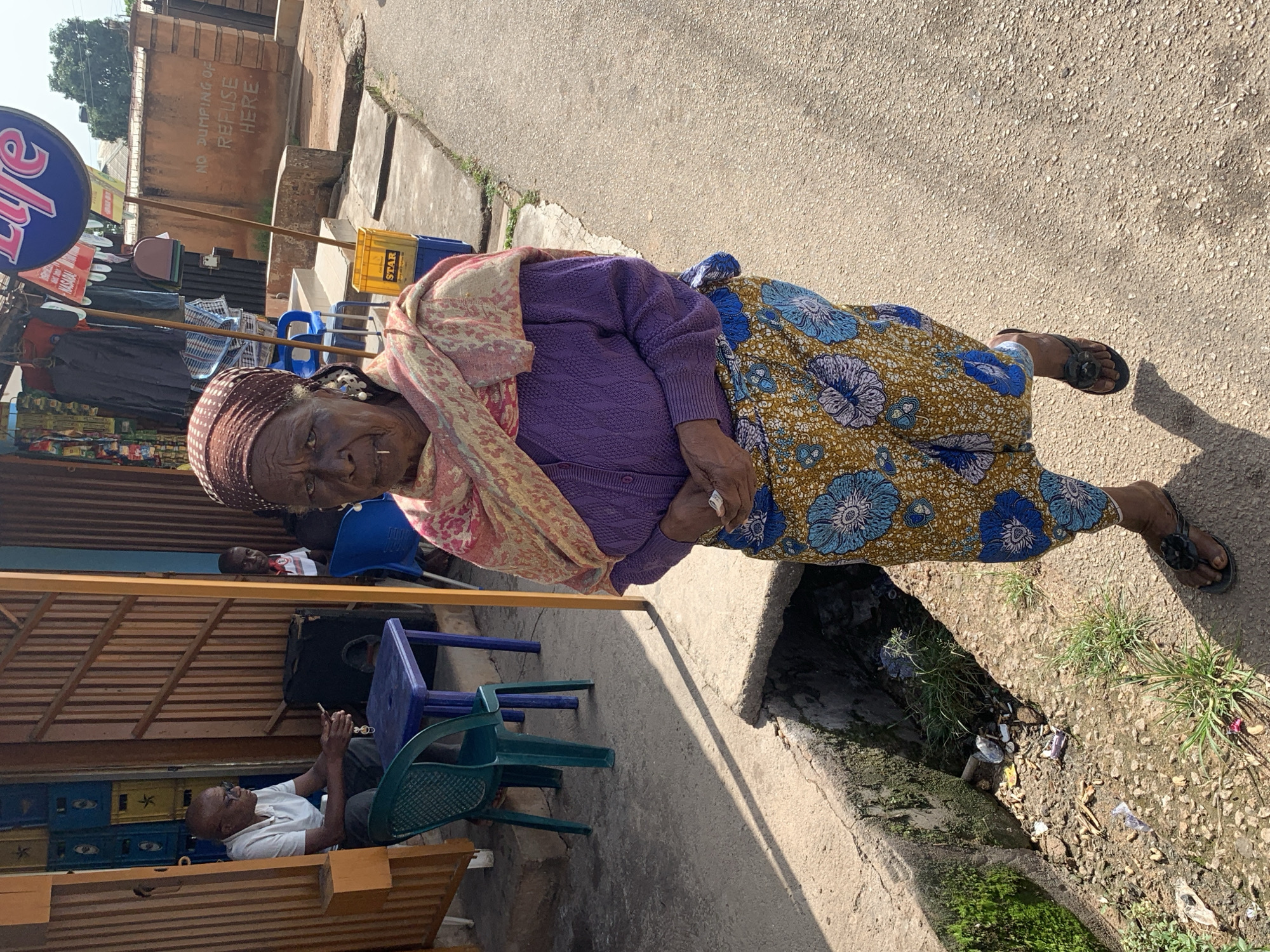 An elderly African woman with a headscarf and traditional clothing stands in the foreground, looking towards the camera. She has a small white object protruding from her mouth. She is wearing a purple knitted cardigan over a patterned dress. To her left, a man in a white polo shirt and glasses is seated at a small blue table, holding a phone. Behind him, a green plastic chair and more blue plastic chairs are visible. The scene appears to be in an outdoor market or shop area, with various items displayed on shelves in the background. A sign with the word "LIFE" in large letters is visible above the shop. The ground is paved with concrete and asphalt, and there is a drainage ditch to the left of the woman, containing debris and vegetation. The environment suggests a warm climate. The location is identified as Jos, Nigeria.