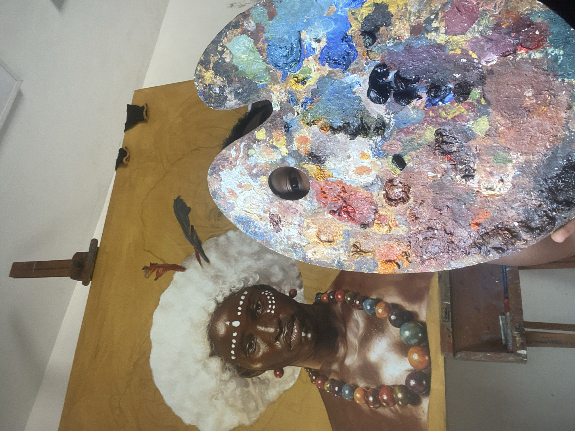 This media file presents an art studio scene, likely located in Ado, Nigeria. The primary subjects are a large canvas painting on an easel and a heavily-used artist's palette.
The painting features a portrait of an individual with dark skin, whose face is decorated with white dot patterns. The subject wears a voluminous white, fluffy headpiece adorned with small black and reddish feather-like elements, and a multi-colored beaded necklace. The person in the portrait exhibits a calm, slightly smiling expression. The painting's background is a warm, earthy golden-brown hue.
In the foreground, an oval wooden artist's palette, held by a partially visible hand, is laden with a thick accumulation of various oil or acrylic paints. Hues of vibrant blues, greens, yellows, reds, browns, black, and white are prominently displayed on its surface, indicating active use. The canvas is supported by a wooden easel, parts of which are visible on the left and lower right of the frame. The setting appears to be an indoor space with plain white walls. The image captures a moment of artistic activity, showcasing the tools and the creation process. There is no visible text in the image.