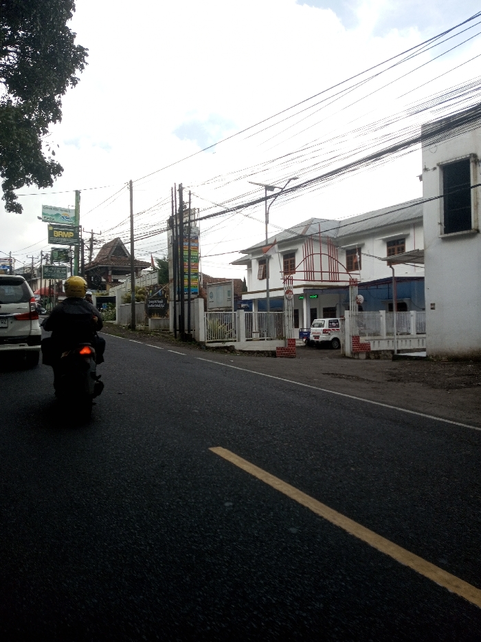 This image captures a daytime street scene, reportedly in the city of Ura-Dozhi, Bhutan, under an overcast sky. The foreground is dominated by a dark asphalt road with a solid yellow line visible on its right side. A person wearing a yellow helmet and dark jacket is riding a dark-colored scooter or motorcycle away from the viewer on the left side of the road, its red taillight illuminated. To the left of the scooter, the rear section of a white SUV is partially visible.
On the right side of the road, a white, two-story building with a gray roof and brown-framed windows stands behind a white fence with red brick pillars. A distinctive red metal archway or decorative element is visible above its entrance. A white van is parked within the fenced area in front of this building. A street light with a solar panel is also present on the right side.
The left side of the road features a green tree with dense foliage. Numerous utility poles support a dense network of overhead wires that crisscross the upper frame. Several commercial signs are visible, including a prominent green sign and a tall, multi-panel vertical sign displaying various colorful graphics and text. Behind these signs, a building with a traditional dark, peaked, tiled roof and wooden architectural elements is discernible.