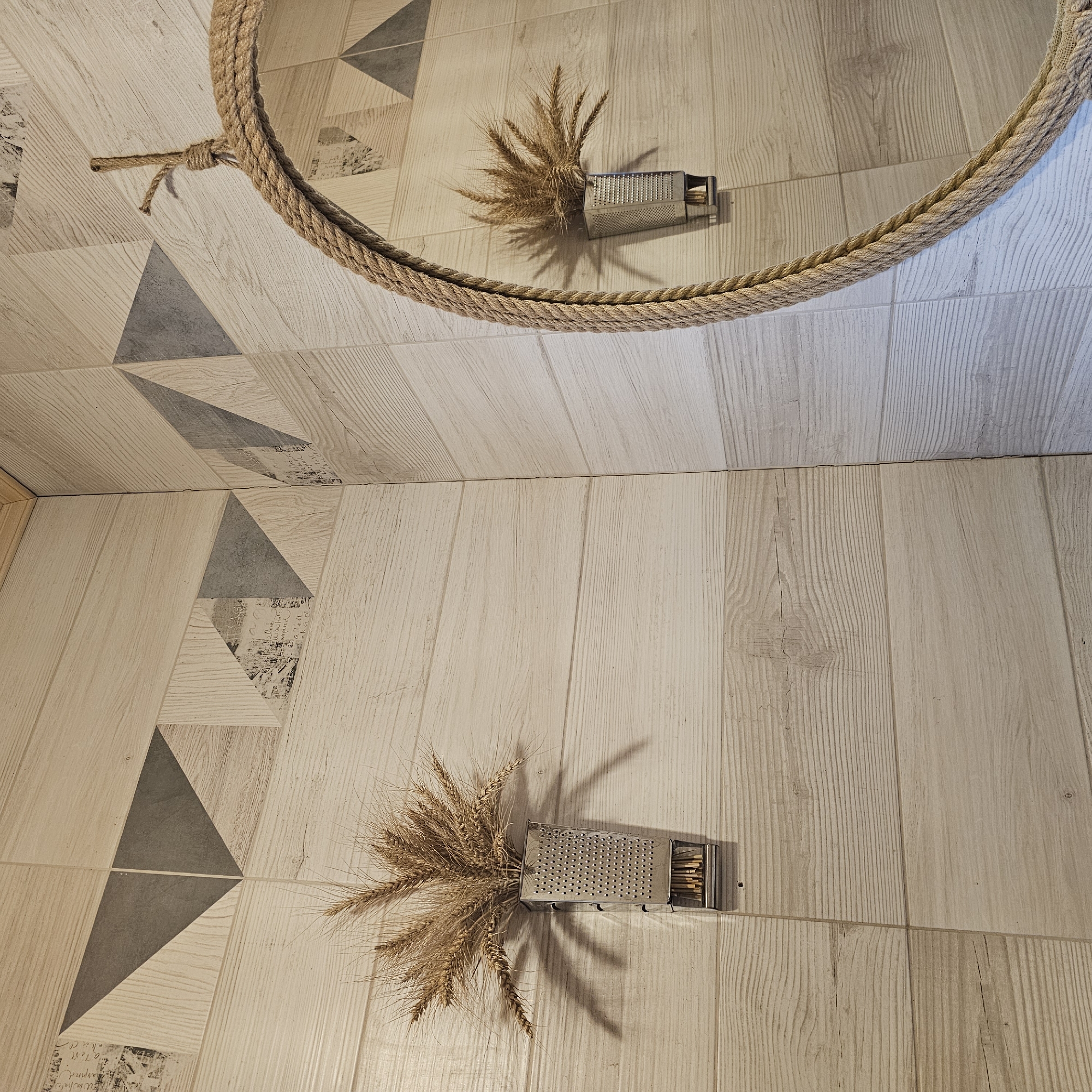 The image shows a close-up view of a tiled floor with a wood-like pattern. A circular mirror with a thick rope frame is partially visible at the top of the image, hanging above the floor. In the reflection of the mirror, a small metal grater is seen filled with dried wheat stalks. On the floor, directly below the mirror's reflection, an identical arrangement of a metal grater filled with dried wheat stalks is placed. The floor tiles feature a geometric pattern of triangles in various shades of beige and grey, some with a subtle script-like texture. The lighting suggests an indoor setting, possibly during the day, casting shadows from the wheat stalks and grater onto the floor. The overall impression is one of rustic, decorative arrangement. No people are visible in the image. The location is identified as Vybolovo, Russia.