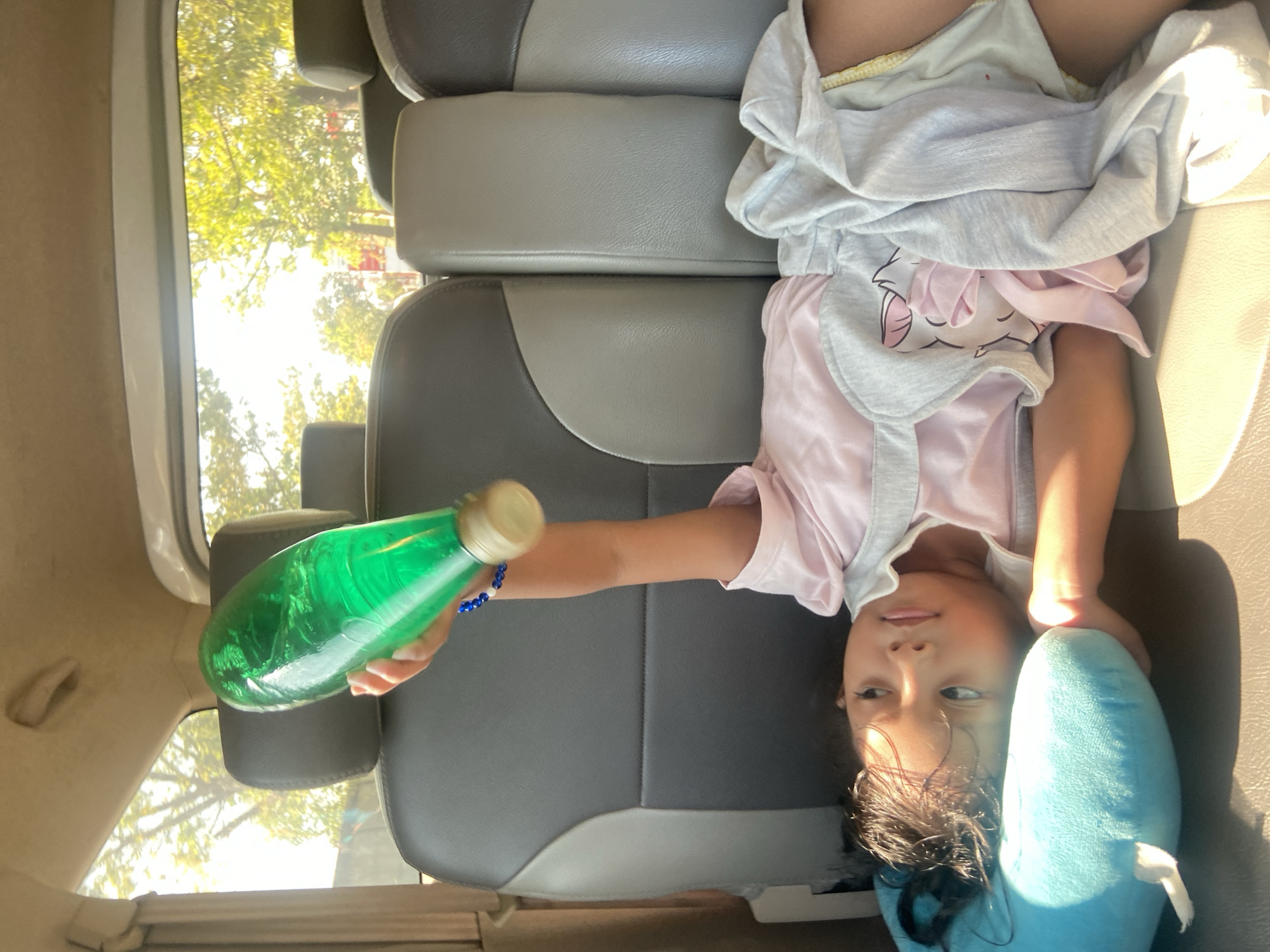 A young girl lies on a car seat with a blue plush toy under her head. She is wearing a pink shirt and grey shorts. Her right arm is raised, holding a green plastic bottle with a blue beaded bracelet on her wrist. The car is likely in motion, with trees and sunlight visible through the windows. The interior of the car appears to be made of grey and beige upholstery.