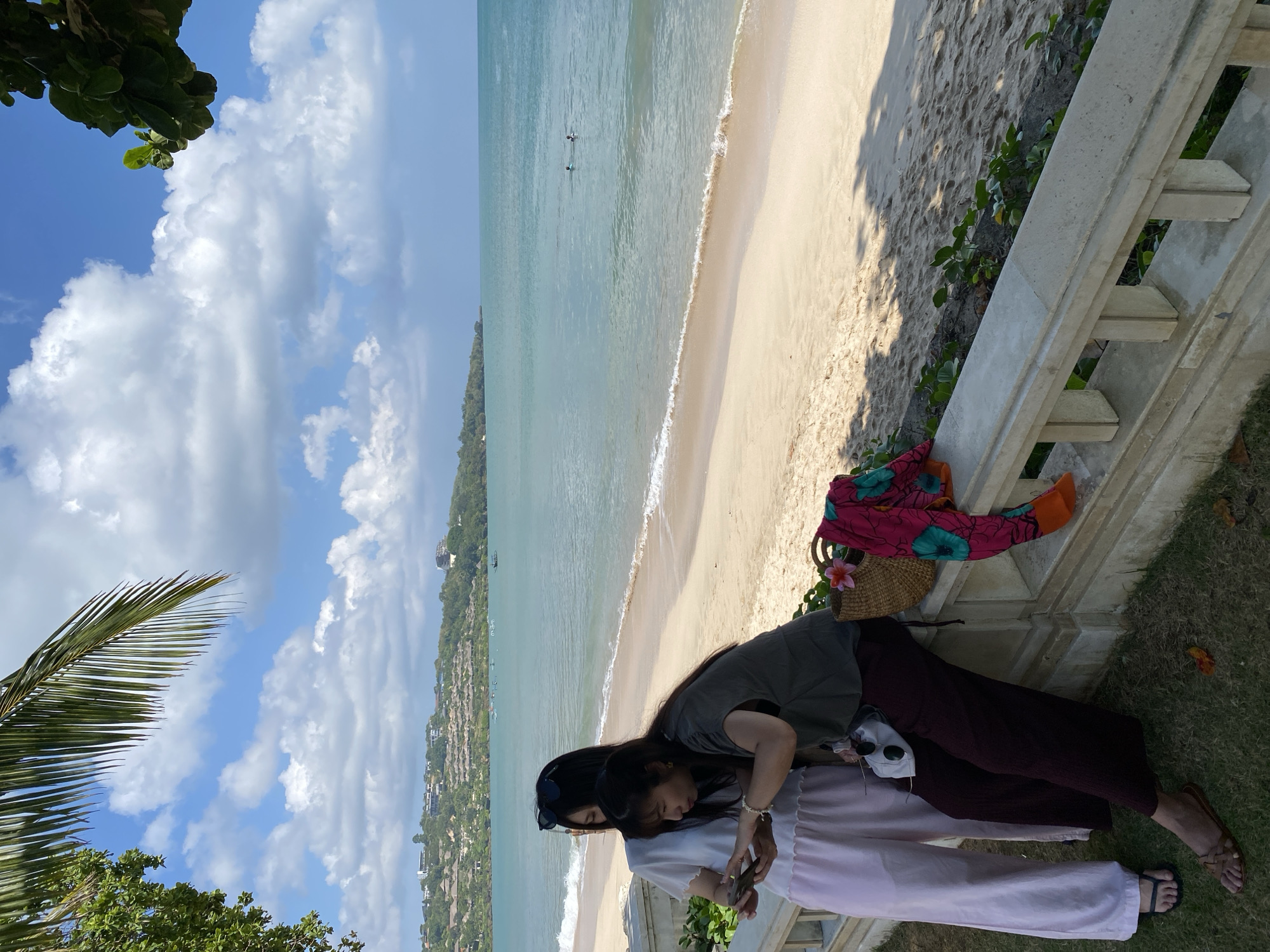 This outdoor image captures a picturesque coastal scene in Kuta Selatan, Indonesia, under a bright, clear sky dotted with white cumulus clouds. The foreground features two women standing on a patch of green grass, adjacent to a concrete railing. One woman, wearing a dark grey top and maroon pants, leans slightly forward, looking down, possibly at a smartphone held by the other woman. The second woman, dressed in a white top and light pink pants, holds a smartphone in her hands. A woven beach bag with a pink flower and a colorful patterned sarong are placed on the grass near the railing beside them.
Beyond the railing, a wide expanse of golden sandy beach stretches along the coastline, meeting the tranquil, turquoise ocean. Gentle waves can be seen breaking at the shore. Further out in the water, two indistinct figures are visible, possibly engaged in swimming or other water activities. The background reveals a lush, green hillside densely populated with numerous buildings, suggesting a developed resort area. Tropical foliage, including a large palm frond, frames the upper left corner of the image. The scene is illuminated by bright daylight, indicating a sunny morning or afternoon. No discernible text is visible in the media file.