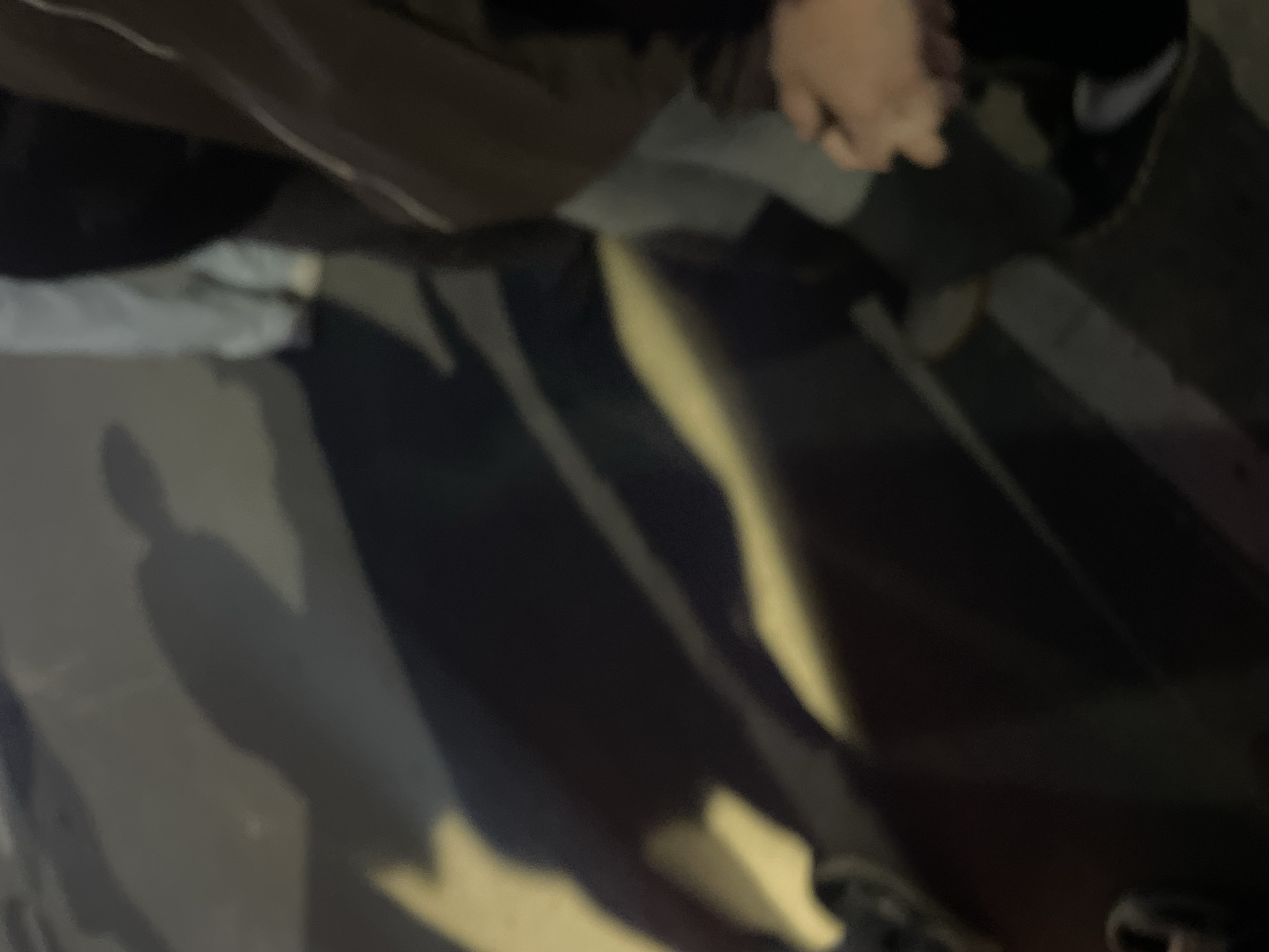The image shows a dimly lit scene that appears to be outdoors at night. A portion of a person is visible from above, with their hand and part of their clothing in the upper center. The ground is a dark, possibly asphalt surface, with long shadows stretching across it. The location is not identifiable as Красные Баки, Russia, based on the visual information provided.