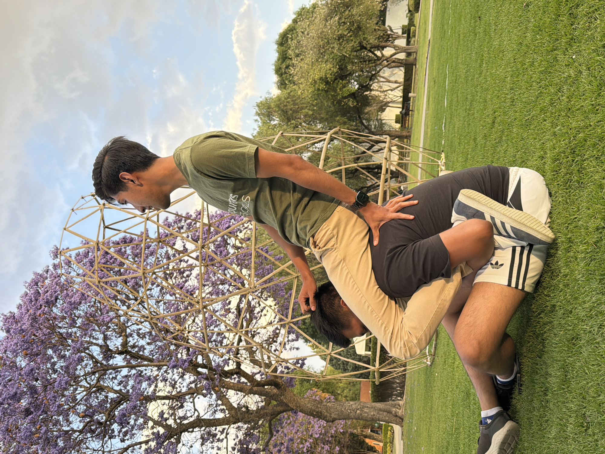 The scene takes place in an outdoor park in San Andrés Cholula, Mexico. Two males are positioned on green grass. One male, dressed in an olive-green t-shirt with partial white text, khaki pants, and a dark smartwatch on his left wrist, is seated on the shoulders of the second male. This upper individual, with dark hair, looks towards the right, with his left hand resting on the head of the male below him. The second male, wearing a black t-shirt and light-colored shorts with dark stripes (Adidas branding visible), sits on the grass with bent legs, supporting the upper individual; his hands are positioned to steady the person above. Behind the two individuals stands a large, light-colored wooden geodesic dome-like structure. To the left of this structure, a large Jacaranda tree with purple blossoms is prominent. Additional green trees line the background, where a paved path and benches are visible in the distance. The sky above is partly cloudy with blue patches.