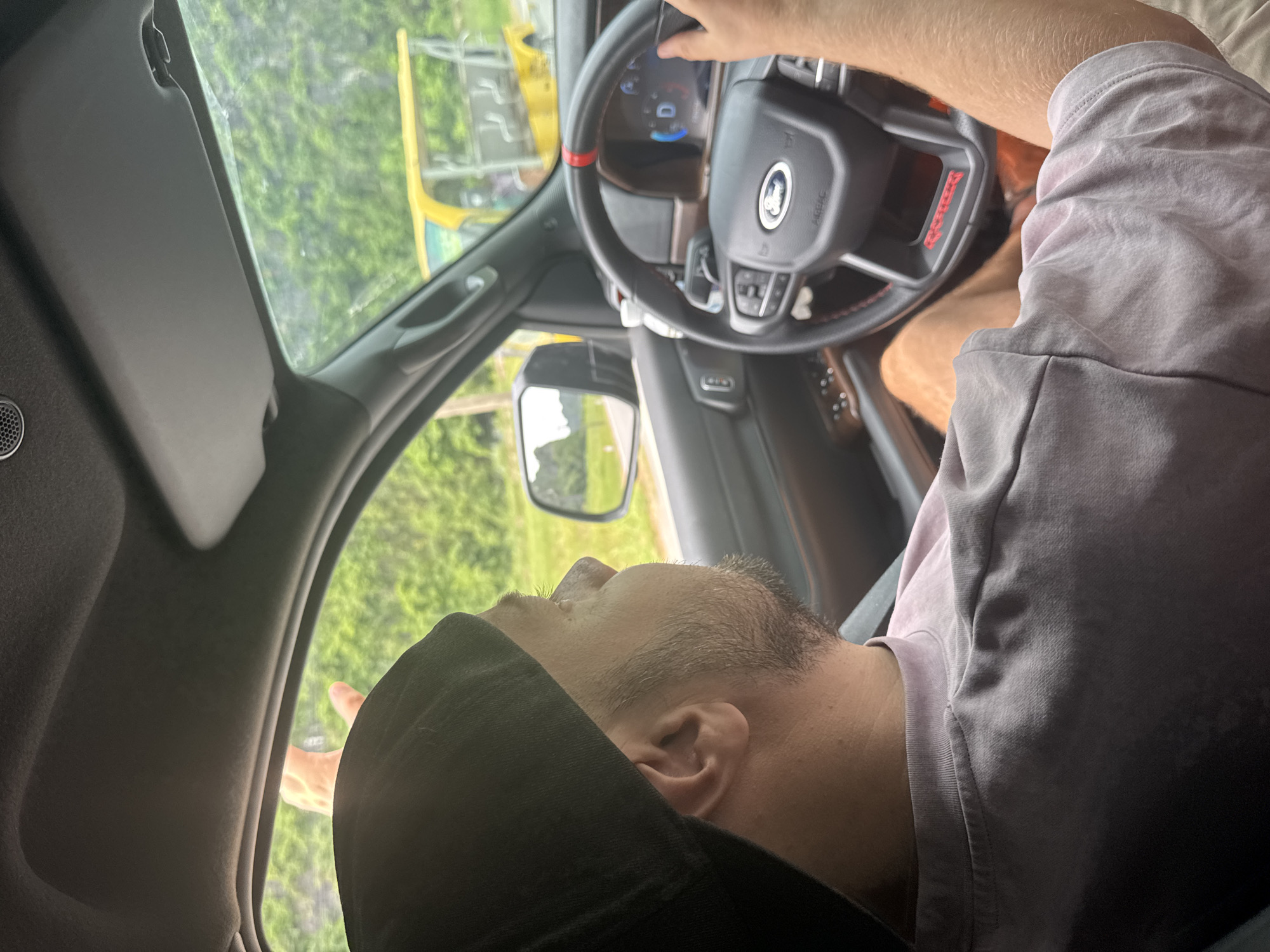 An over-the-shoulder shot from inside a vehicle shows a man driving. The man is wearing a black baseball cap and a light purple t-shirt. He is looking to the left, out the side window. The steering wheel of the car is visible, with the Ford logo at the center and a digital dashboard displaying "D" and "3". Outside the car, a yellow golf cart-like vehicle is visible, along with lush green vegetation and trees. The location is Nam Hoa Lư Ward, Vietnam.
