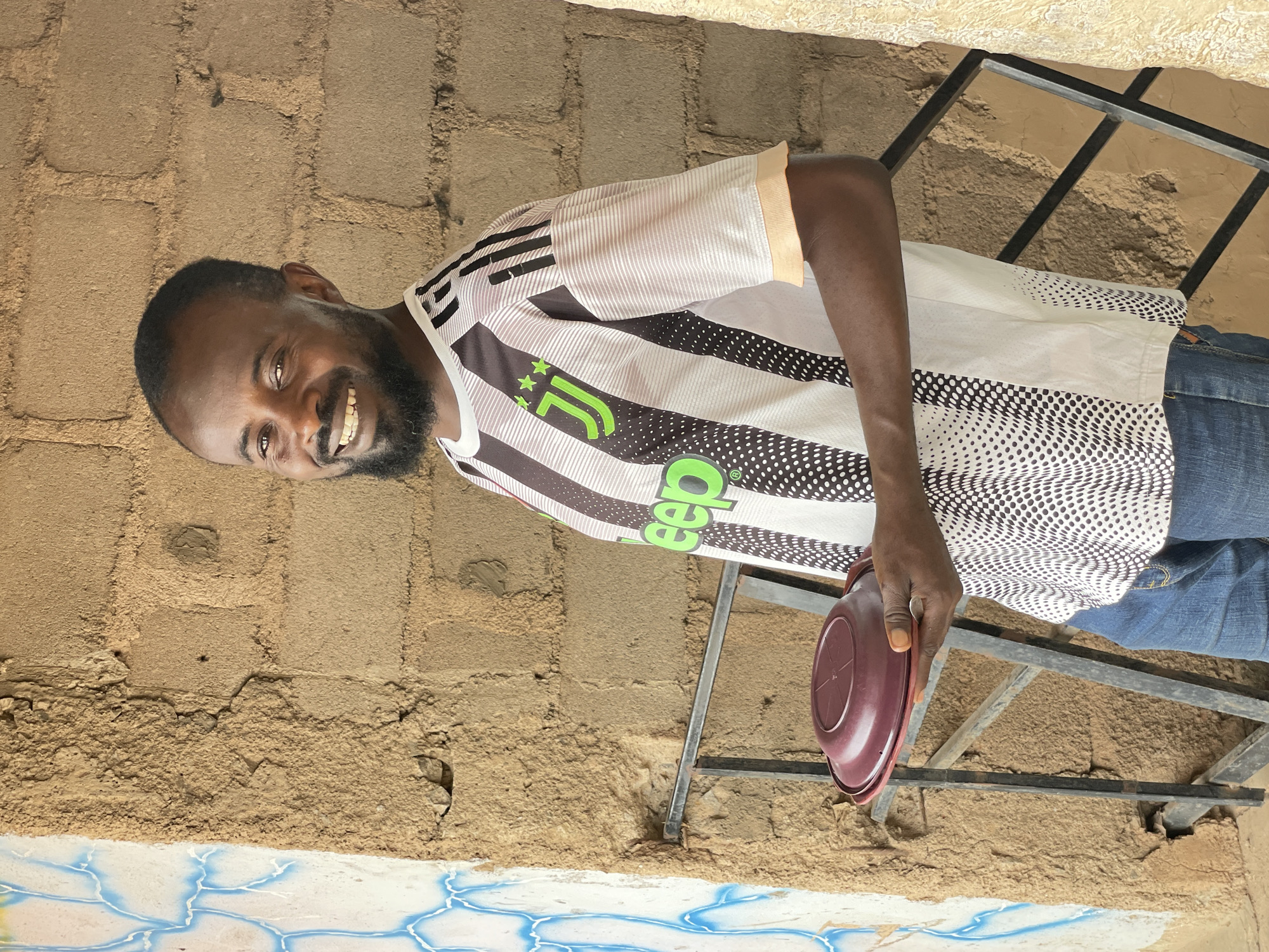 A man is smiling and looking at the camera while holding a burgundy plastic bowl. He is wearing a white jersey with black stripes, and a green Jeep logo. He is standing outdoors near a rough, tan-colored brick wall. The location is assumed to be Bukuru, Nigeria. There is a metal structure, possibly part of a building or a ladder, visible behind him. The lower part of the image shows a white surface with blue and yellow abstract designs.