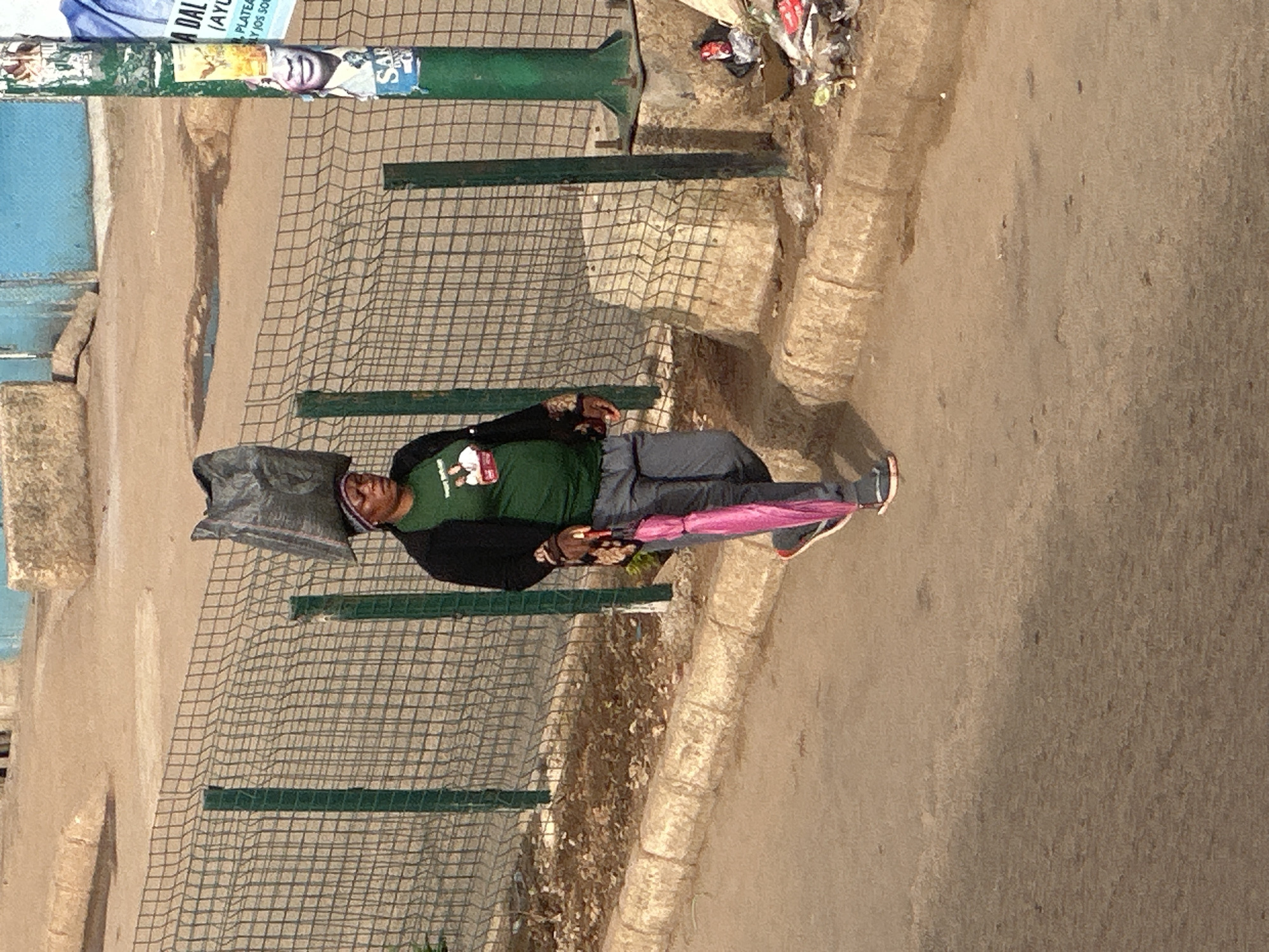 A woman walks along a dusty road in Bukuru, Nigeria, carrying a large black bag on her head. She is wearing a green t-shirt, dark jacket, and grey pants, and holds a closed pink umbrella in her right hand. A wire fence runs alongside the road, with a concrete curb and scattered debris on the other side. In the background, there are some buildings and a light blue wall. The scene is illuminated by natural light, suggesting it is daytime.